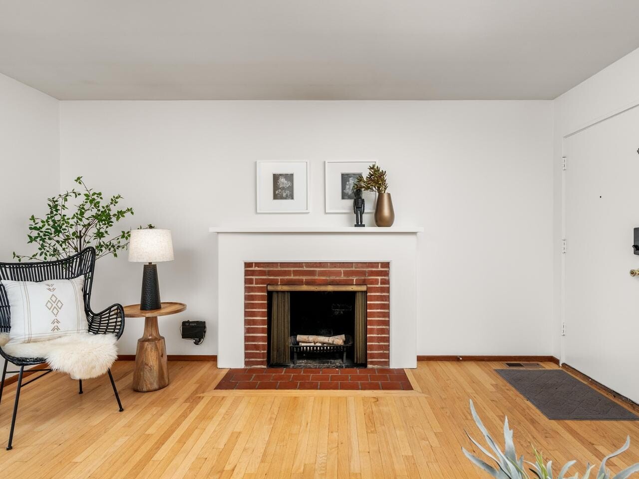 A minimalist living room with a brick fireplace centered on a white wall. Two framed pictures and a vase are on the mantel. A wicker chair with a pillow and throw sits to the left, beside a small wooden table with a lamp. Light wood flooring completes the space.