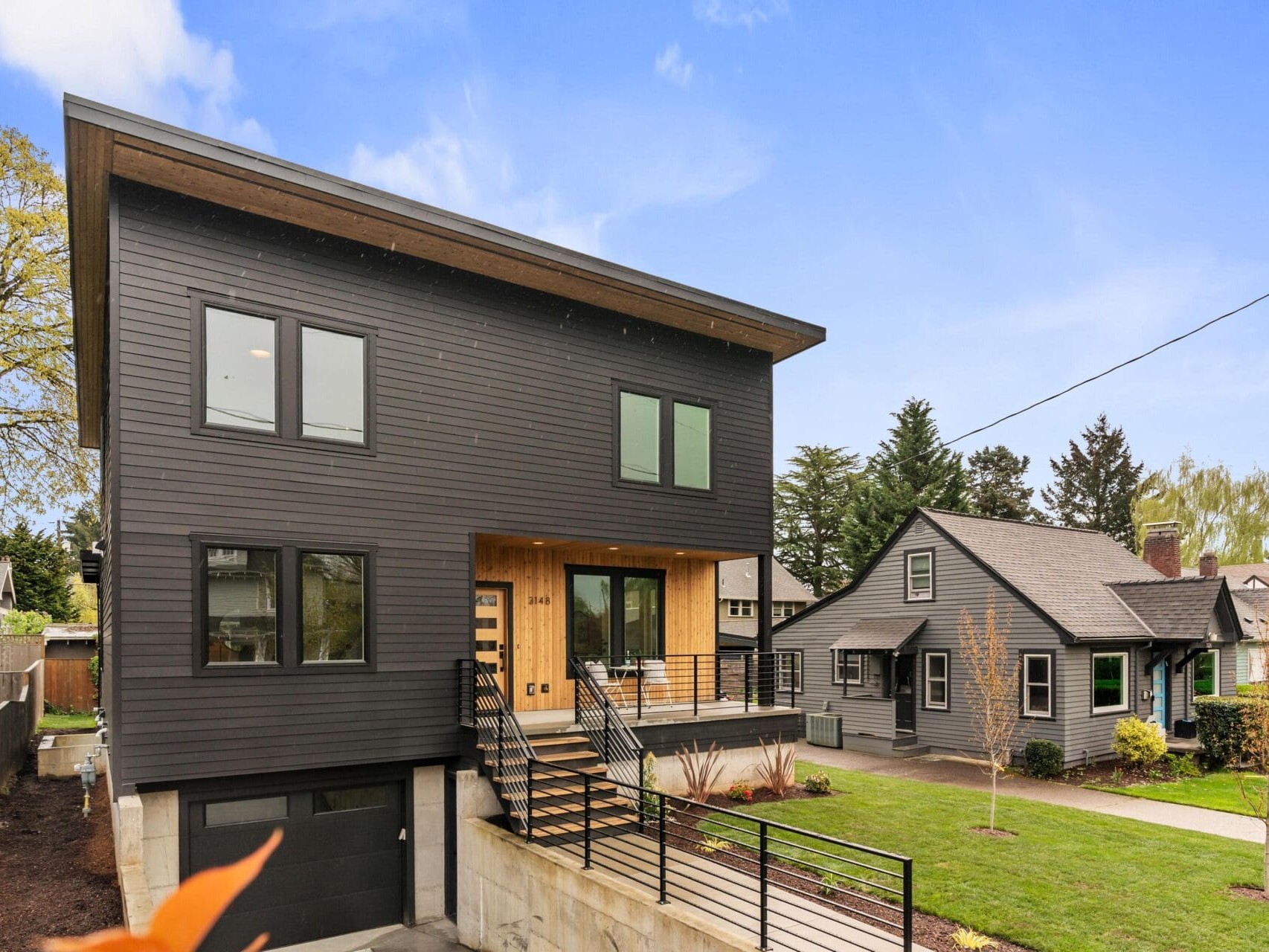 A modern, two-story house with dark siding, large windows, and a slanted roof. It features a raised entrance with stairs and a garage below. A smaller, traditional house with a chimney is next to it. The sky is clear and blue.