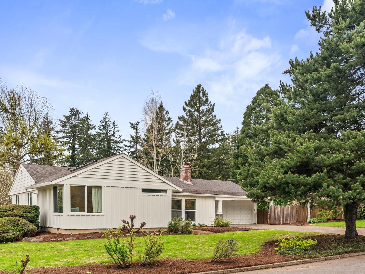 Single-story white house with large windows and a gray roof, surrounded by green lawn, shrubs, and tall trees under a blue sky with scattered clouds. A driveway is visible on the right.