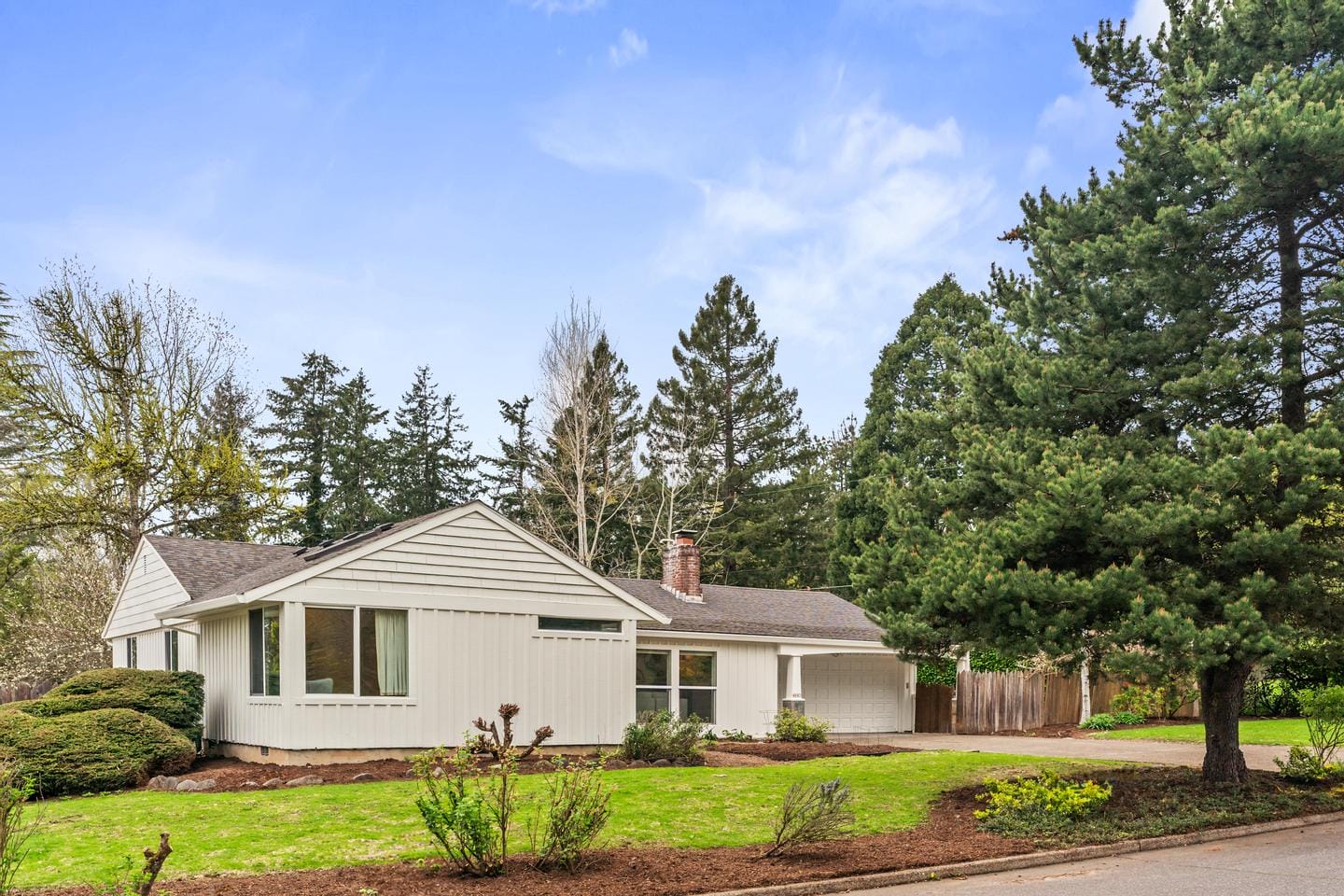 Single-story white house with large windows and a gray roof, surrounded by green lawn, shrubs, and tall trees under a blue sky with scattered clouds. A driveway is visible on the right.