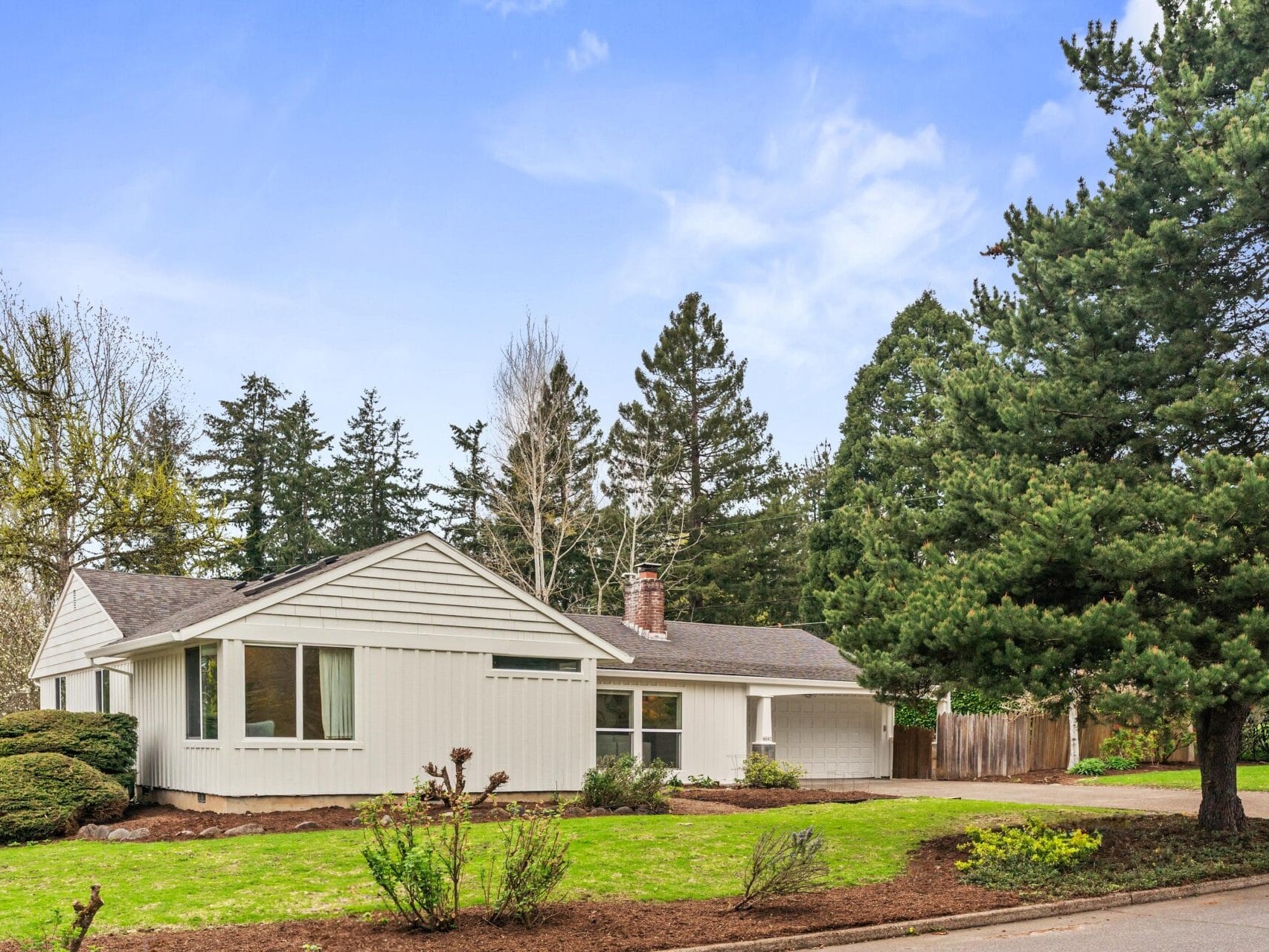 A single-story white house with a gray roof sits on a green lawn, surrounded by tall trees and shrubs under a blue sky with light clouds.
