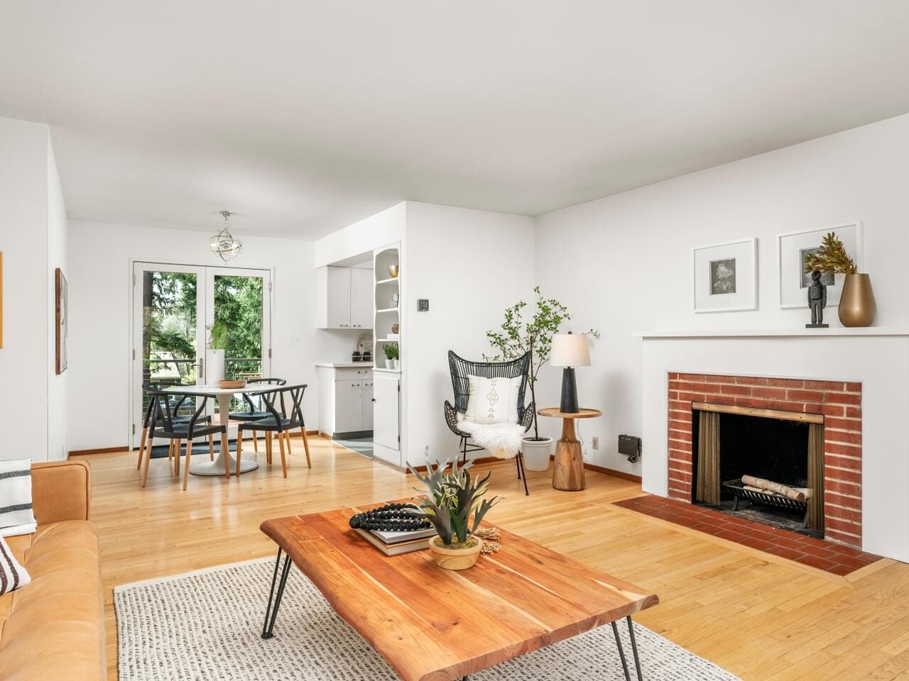 A bright living room with a wooden floor, featuring a brick fireplace, a wooden coffee table, and a cozy sofa. A dining area is visible in the background next to the kitchen. Large windows offer a view of greenery outside. Minimalist decor accents the room.