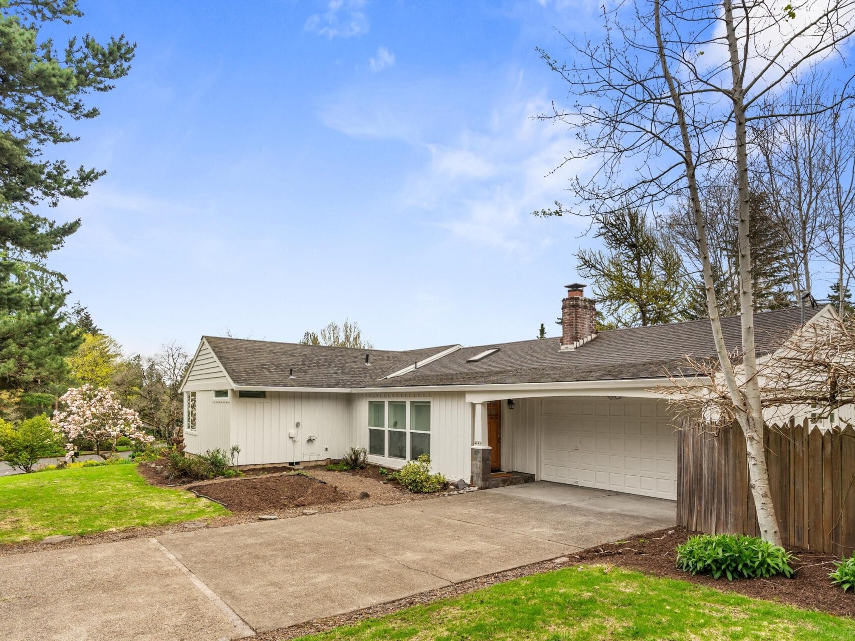 Single-story white house with attached garage, gray roof, and large driveway. The front yard has green grass, trees, and shrubs, with blue sky and scattered clouds in the background.