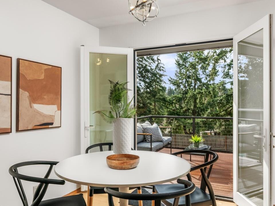 A dining area with a round white table surrounded by four black chairs. Two abstract paintings hang on the white wall. French doors open to a deck with outdoor seating and a view of green trees. A modern light fixture hangs above.