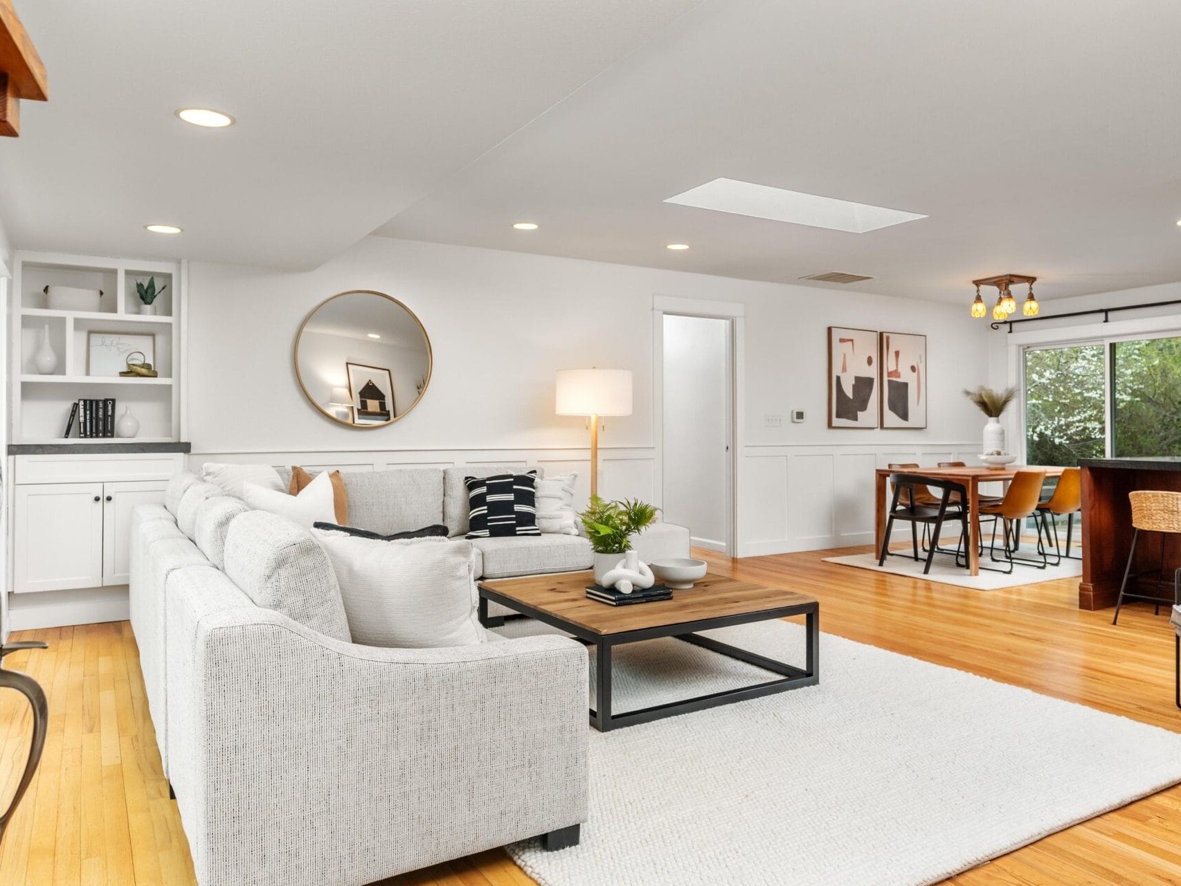 A modern, open-concept living and dining area with white walls, a light gray sectional sofa, wooden floors, and minimalist decor. A round mirror, framed art, and large windows create a bright, airy atmosphere.