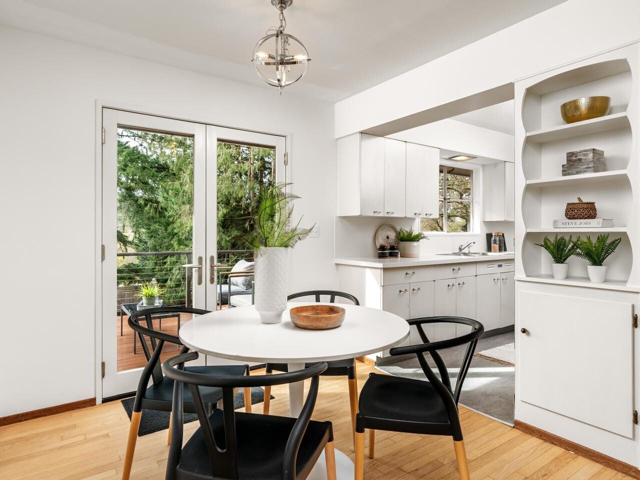 A modern dining area with a round white table and four black chairs. A large plant sits in the center. French doors open to a balcony with greenery outside. A kitchen with white cabinets is visible to the right, featuring shelves with plants and decor.