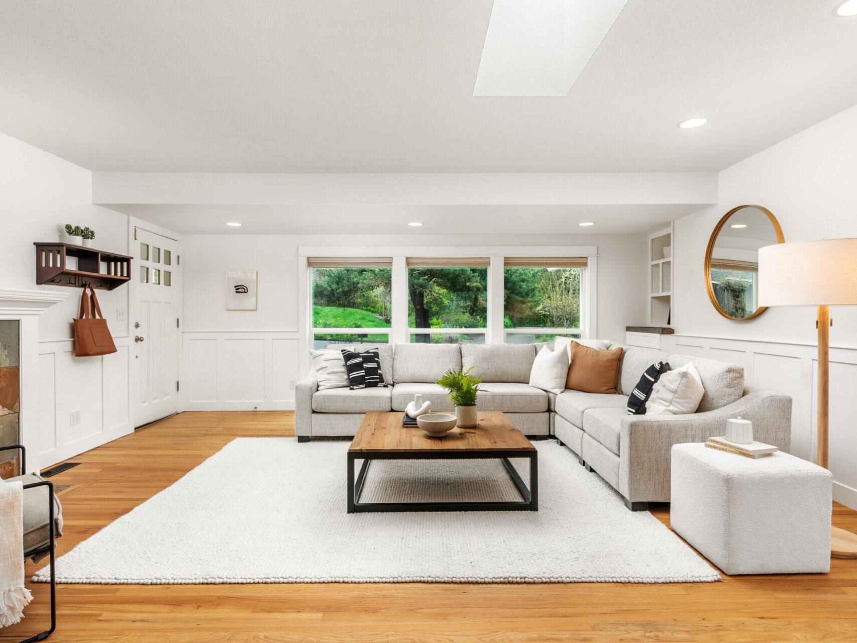 Bright living room with large windows, a beige sectional sofa, a wooden coffee table on a white rug, a floor lamp, a round wall mirror, and hardwood floors. Green trees are visible outside.