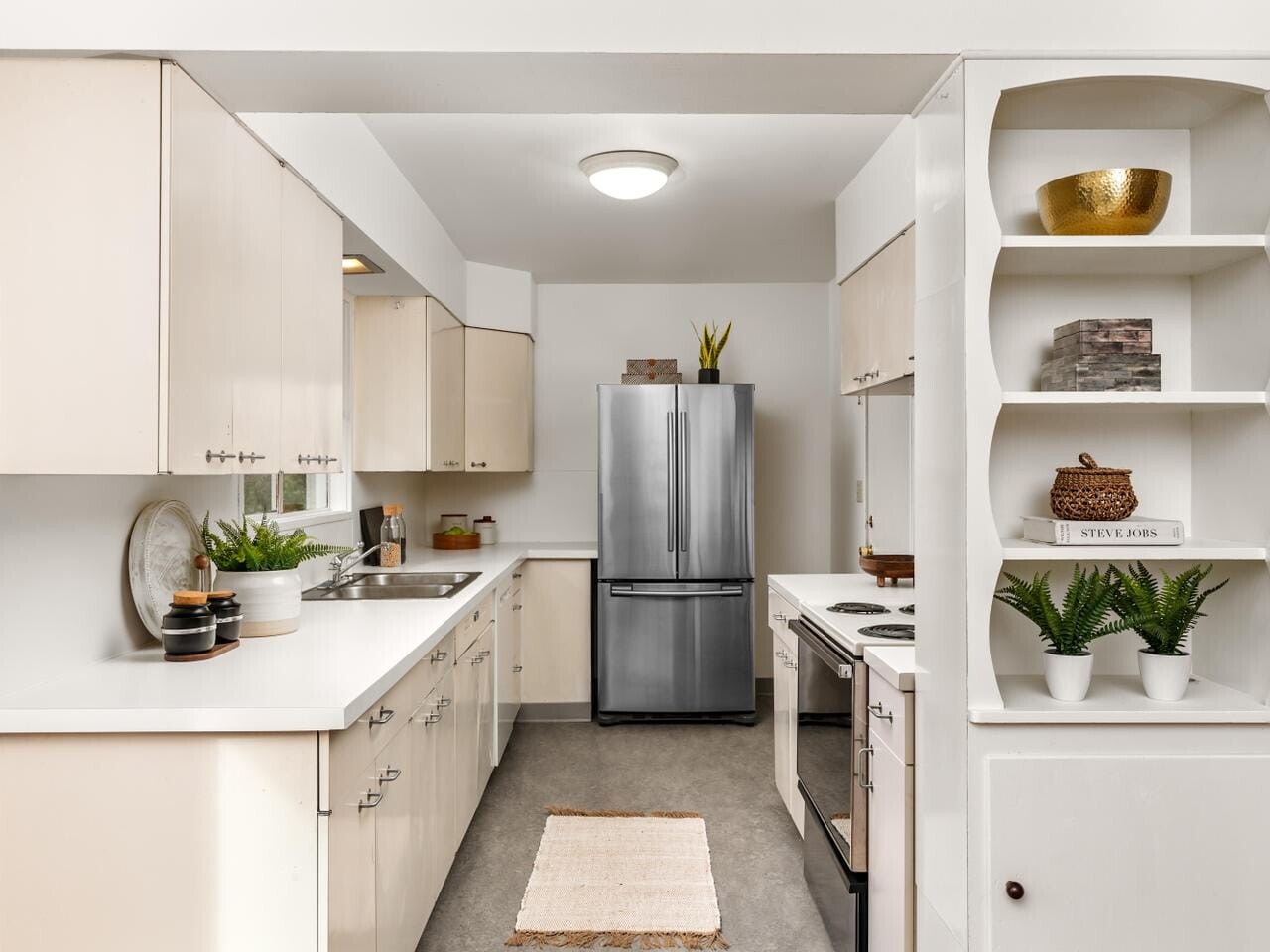 A modern kitchen with white cabinetry and silver appliances. The room features a double-door refrigerator, small plants, and decorative items on open shelves. A beige rug lies on the grey floor, and a round ceiling light illuminates the space.