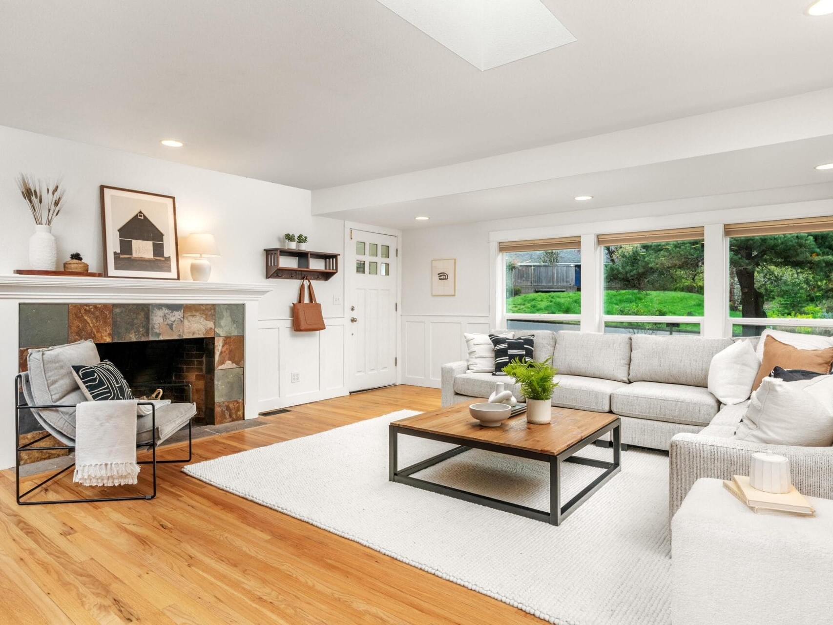 Bright, modern living room with a gray sectional sofa, wooden coffee table, fireplace with slate surround, armchair, hardwood floors, large windows, and neutral decor accents.