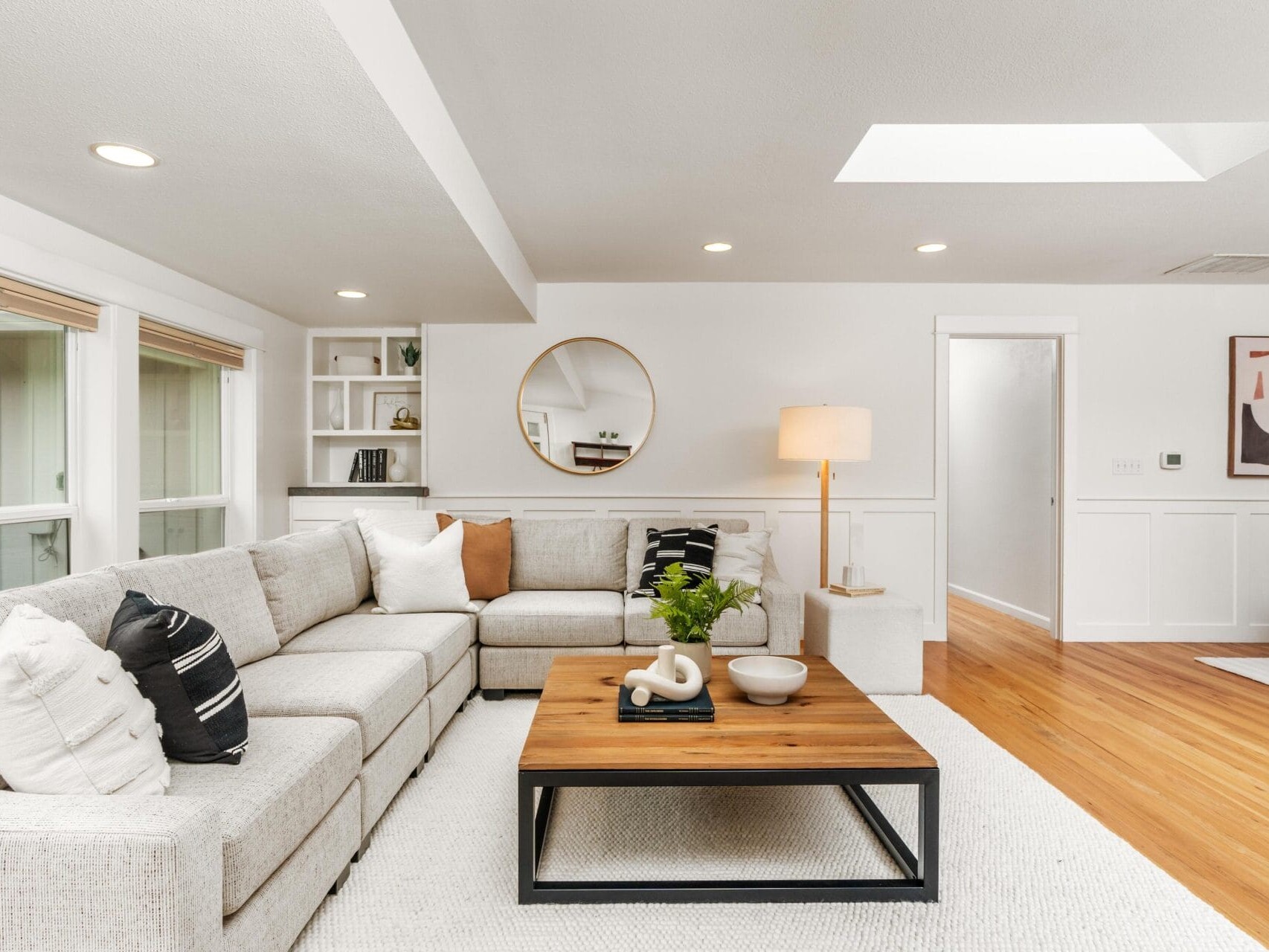 A modern living room with a light gray sectional sofa, neutral pillows, a wooden coffee table with decor, a round mirror, a floor lamp, built-in shelves, and hardwood floors under natural and recessed lighting.