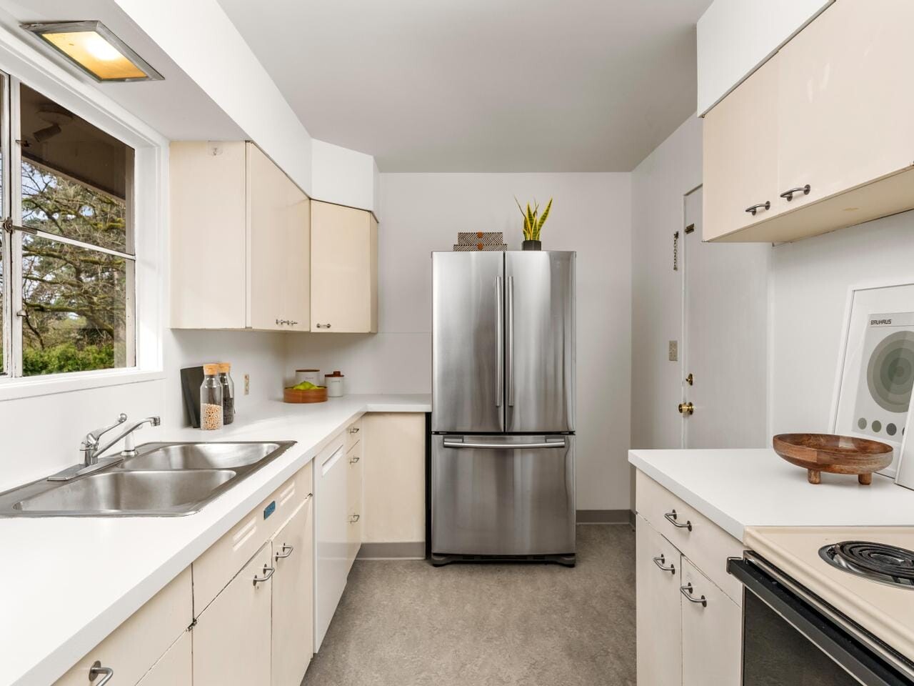 A modern kitchen with white cabinets, a stainless steel refrigerator, and a double sink below a window. The countertops are white with assorted kitchen items, and theres a small potted plant on the fridge. An electric stove is on the right.