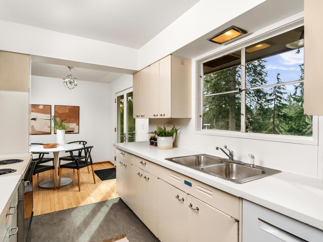 A bright kitchen with light cabinetry, a double sink, and view of trees through the window. To the left, a table with chairs is visible in the dining area. Decor includes plants and framed art. Warm lighting enhances the wooden floor.