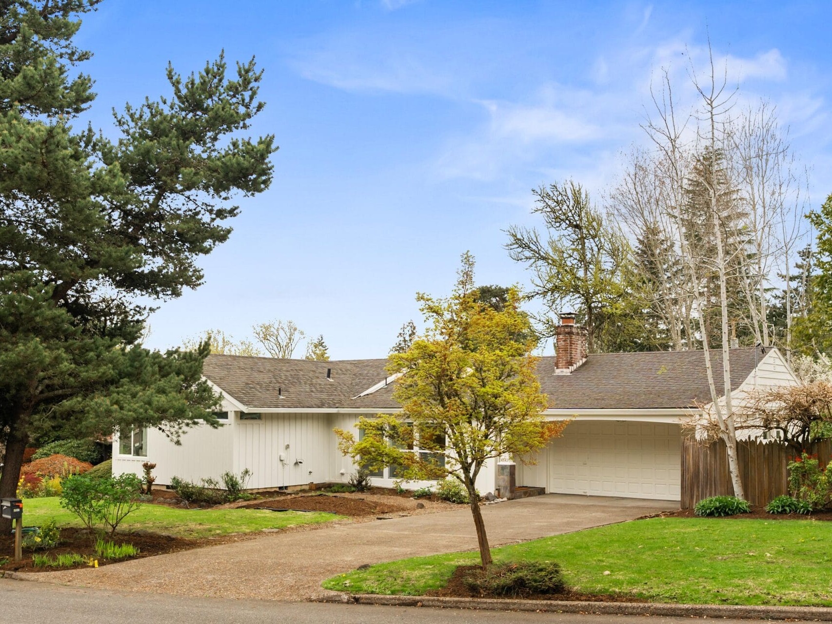 A single-story white house with a gray roof and attached garage sits on a well-kept lawn with trees and shrubs, under a blue sky on a suburban street.