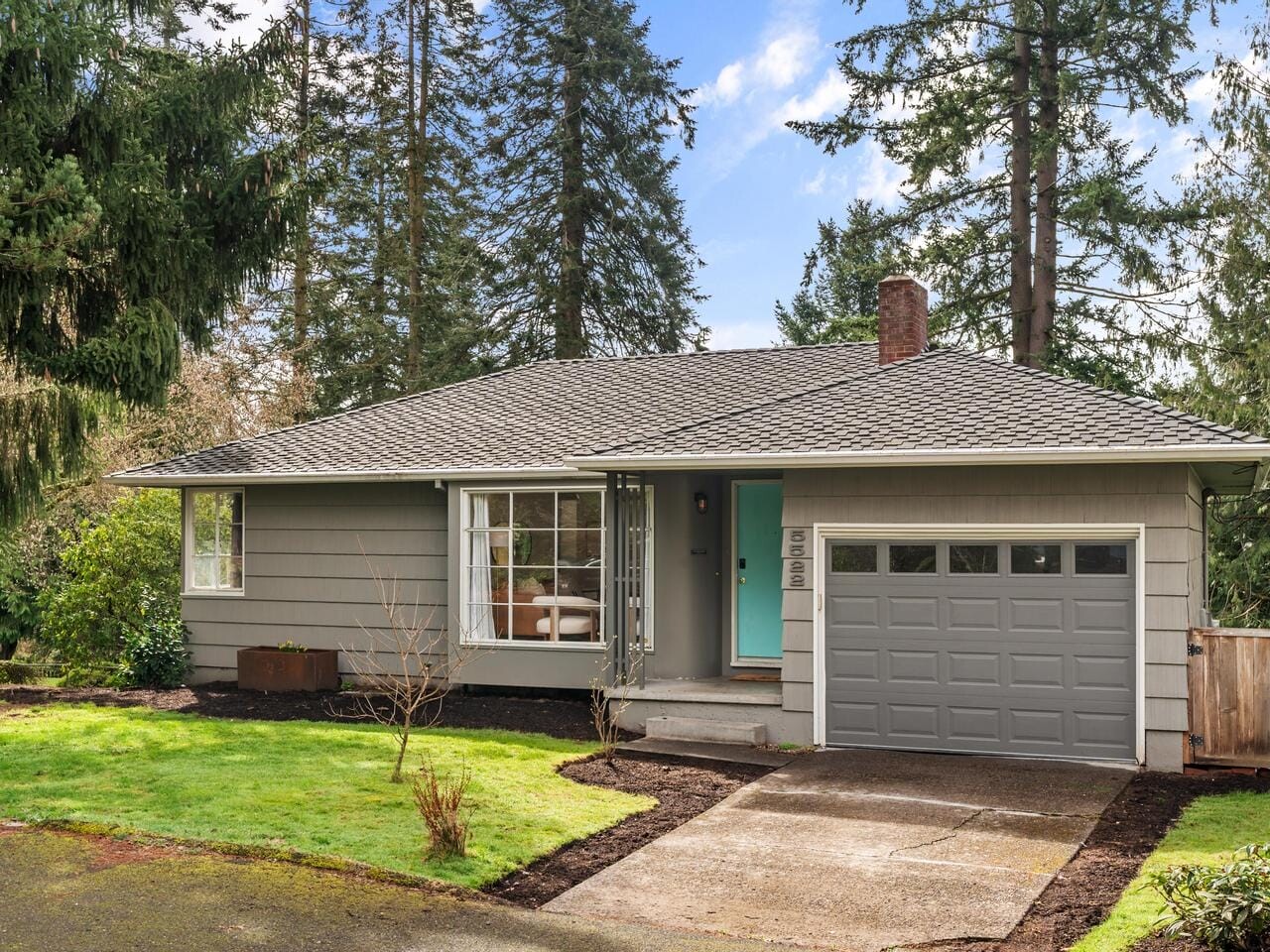 A small, single-story gray house with a garage and a teal front door is surrounded by tall pine trees. The yard has green grass and a few plants. The sky is partially cloudy.