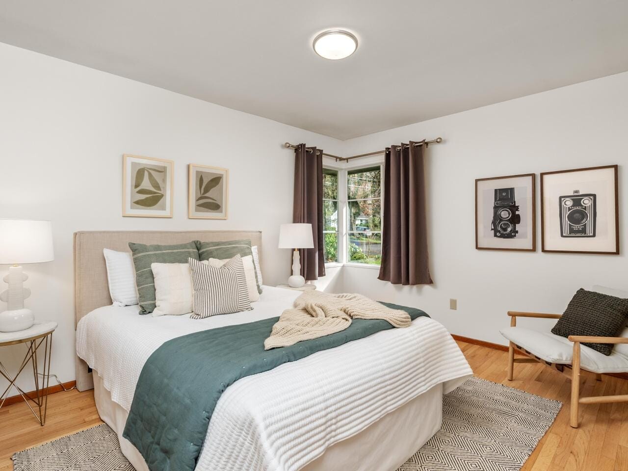 A cozy bedroom with a bed covered in white and green bedding, a nightstand with a lamp, dark curtains on a corner window, and stylish prints on the walls. A wooden chair with a cushion sits on a textured rug over hardwood floors.