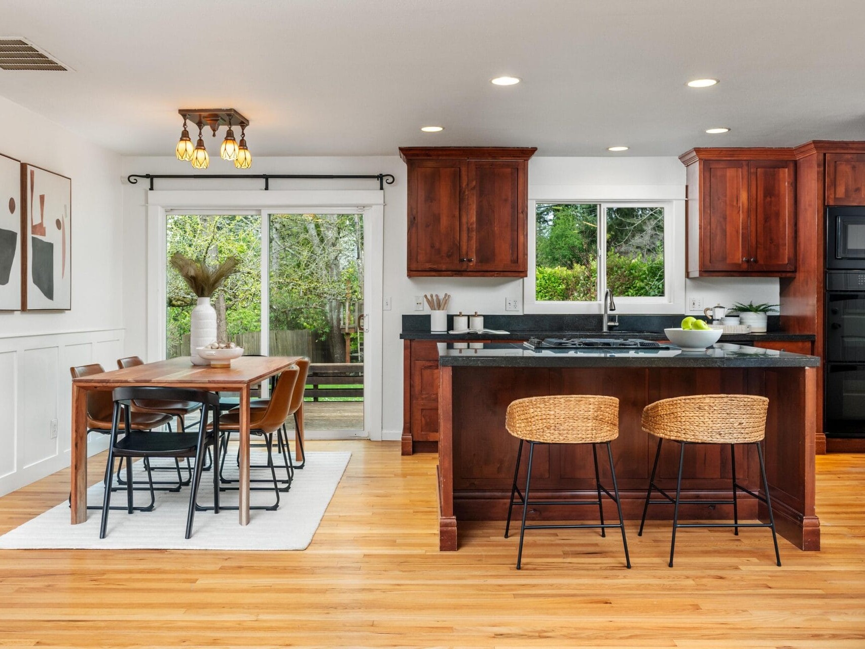 Modern kitchen and dining area with wooden floors, dark wood cabinets, black countertops, a kitchen island with two wicker stools, and a dining table with six chairs near sliding glass doors to a green backyard.