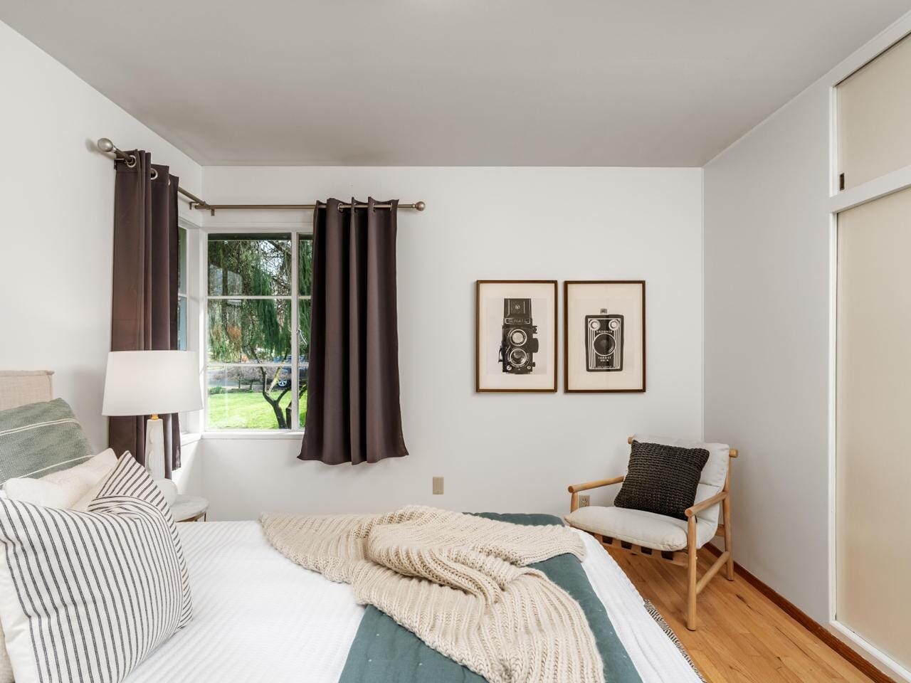 A cozy bedroom with a bed adorned with a green and white striped blanket, decorative pillows, and a beige throw. A wooden chair with a cushion sits by the window, which has dark curtains. Two framed pictures hang on the wall.