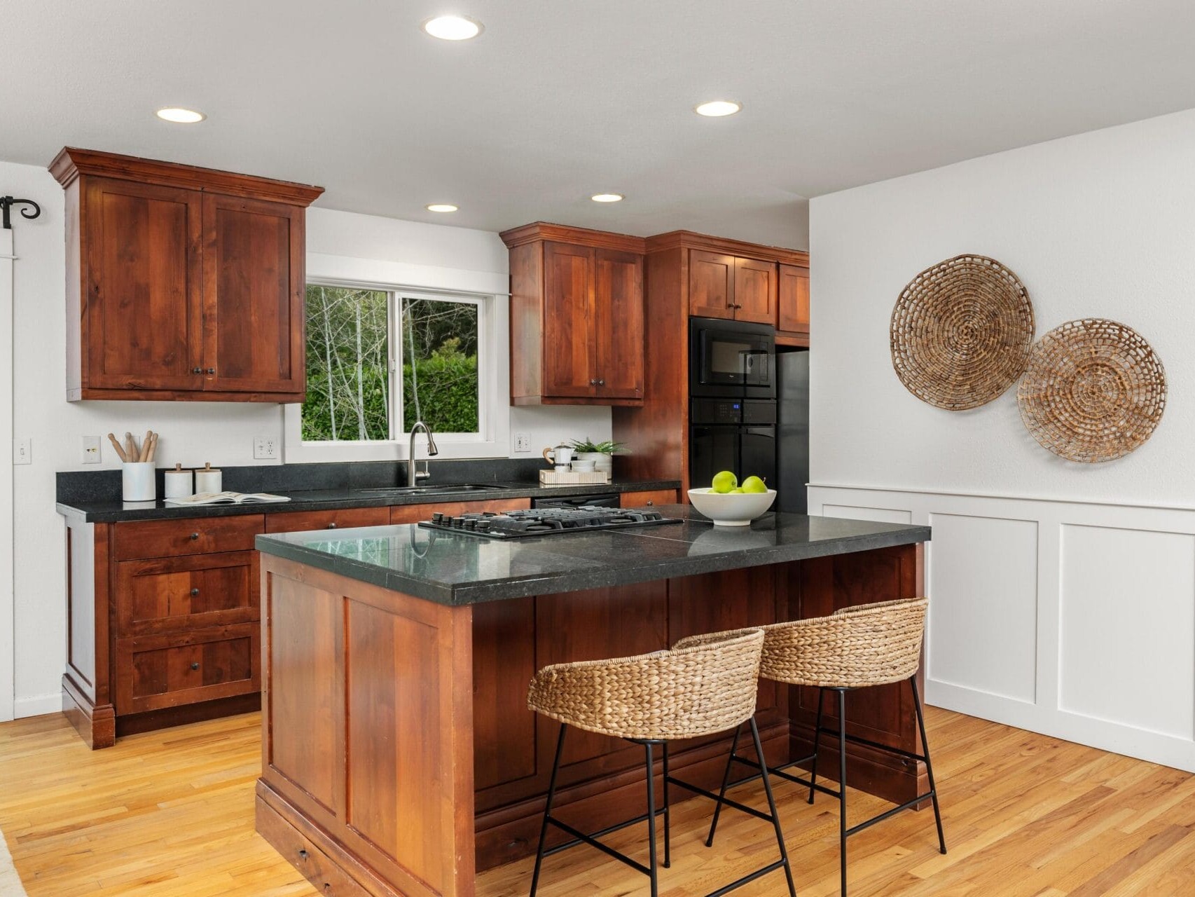 Modern kitchen with dark wood cabinets, black countertops, an island with two wicker barstools, built-in black appliances, wood flooring, and woven wall decor on a white wall. A window overlooks greenery outside.