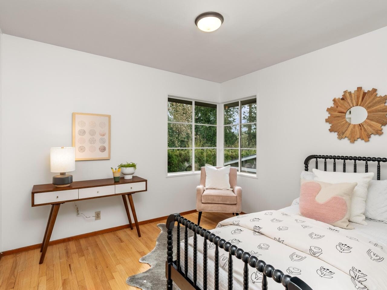 A bright bedroom with hardwood floors features a bed with floral bedding, a pink armchair by the window, a wooden sunburst wall decor, and a mid-century modern desk with a lamp and small plant. Large windows let in natural light.
