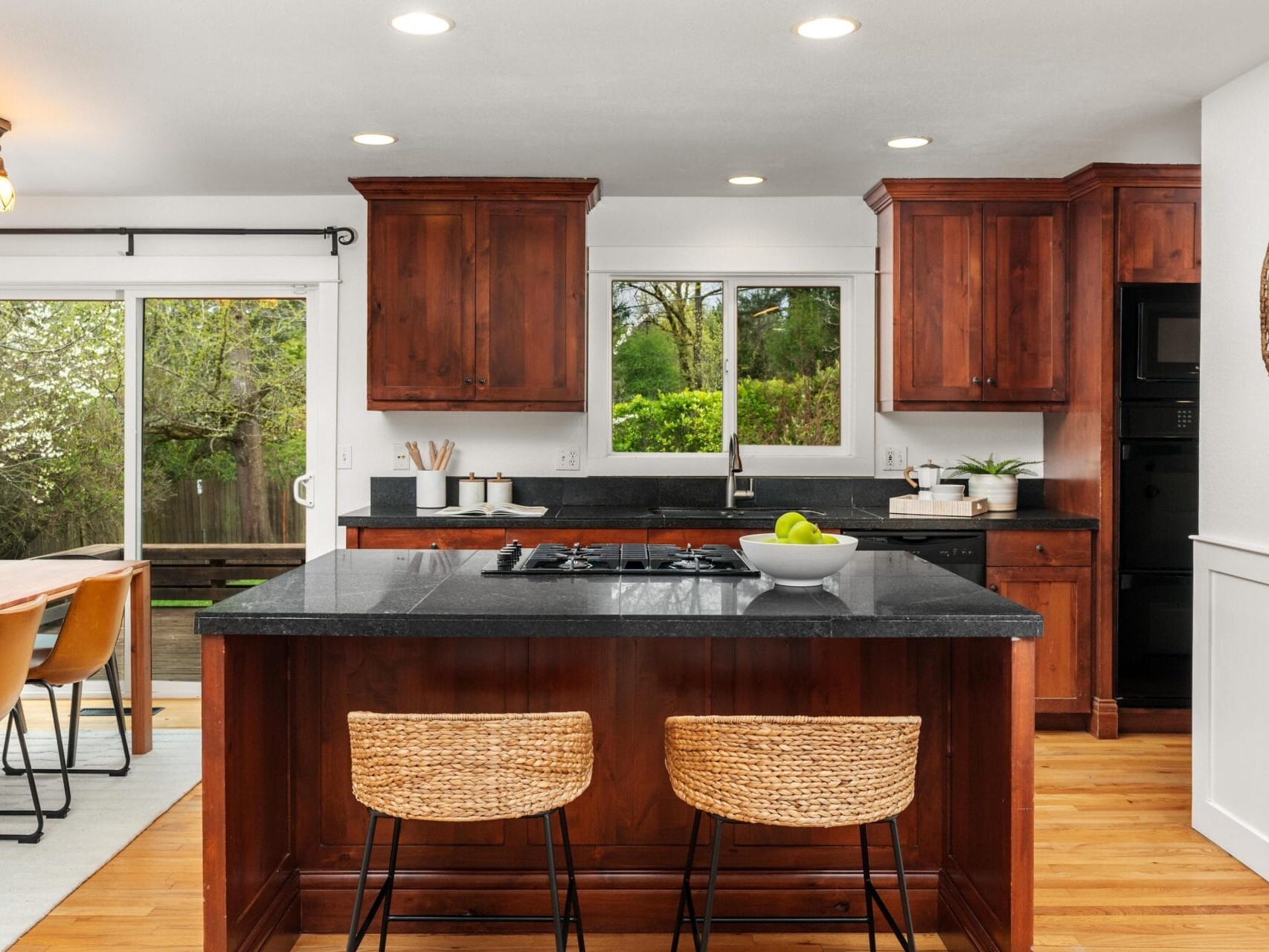 Modern kitchen with dark wood cabinets, black countertops, a center island with wicker stools, and a bowl of green apples. Large window and sliding glass door offer views of greenery outside. Dining area visible to the left.