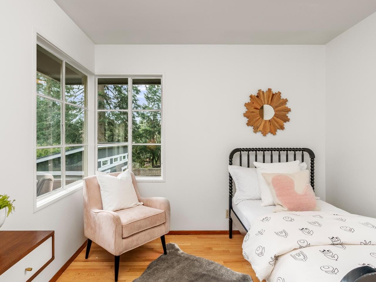 A cozy bedroom with a single bed featuring floral-patterned bedding, a blush armchair, a wooden dresser with a white vase and plant, and a sunburst mirror on the wall. A large window reveals a view of trees outside.
