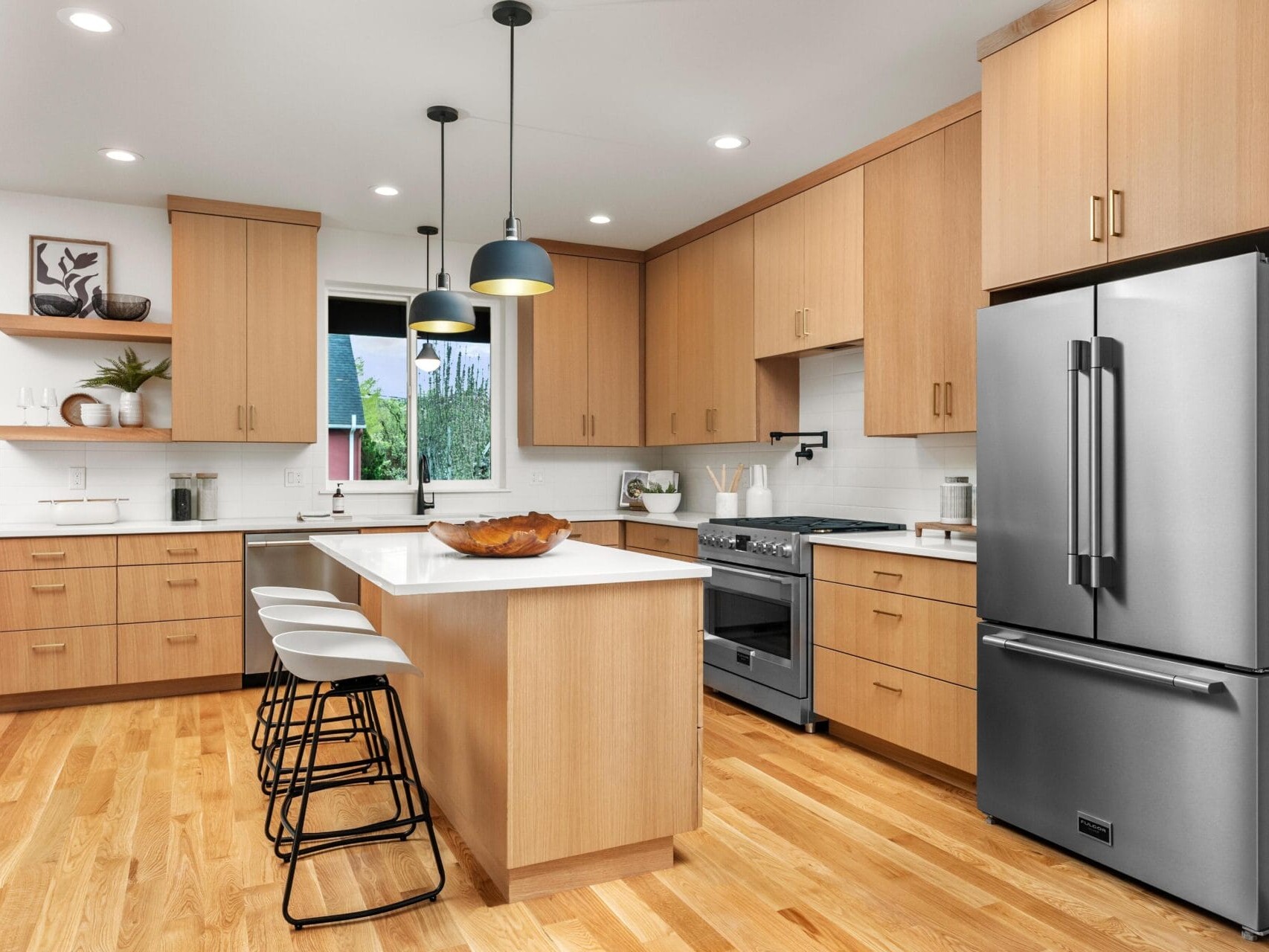 A modern kitchen with wooden cabinets, a large stainless steel refrigerator, and a central island with three bar stools. Pendant lights hang above the island, and open shelves display plants and decor. Hardwood floors match the cabinetry.