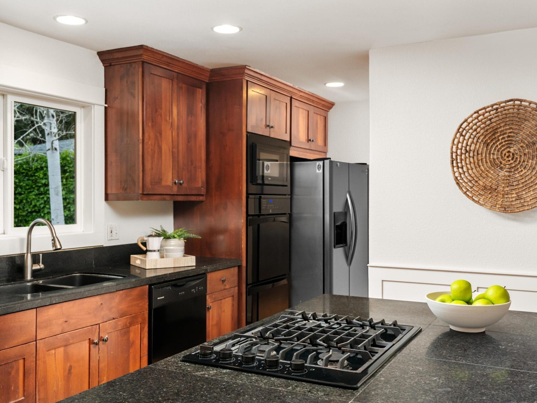 Modern kitchen with dark granite countertops, wooden cabinets, a gas stove, double wall oven, stainless steel fridge, sink under a window, and decorative woven wall art. A bowl of green apples sits on the island.