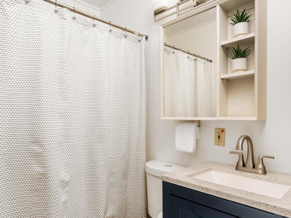 A modern bathroom with a navy blue vanity and beige countertop. Theres a white toilet and a shower with a white, textured curtain. Above the mirror is a light fixture with three bulbs, and shelves hold decorative plants.