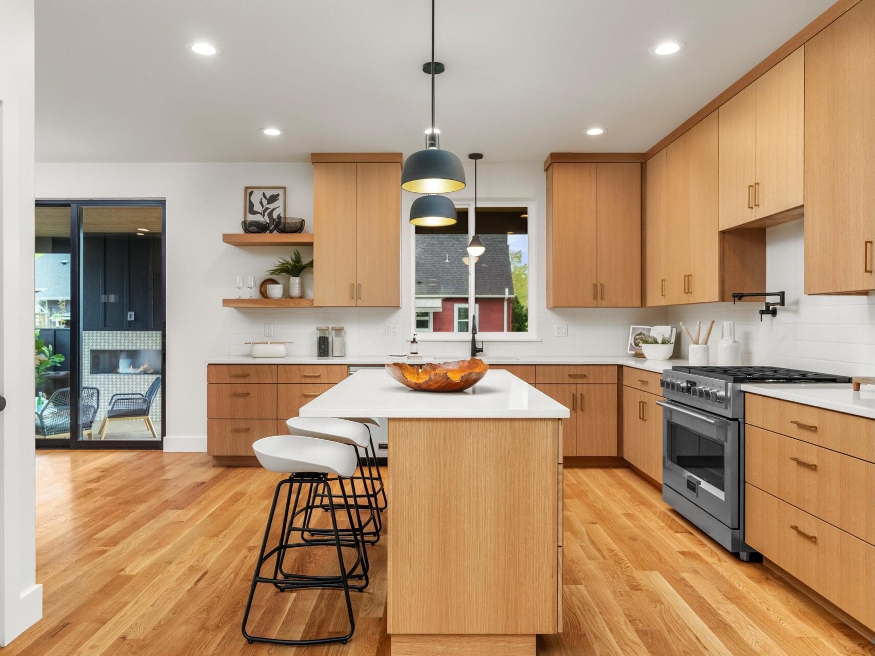 Modern kitchen with wooden cabinets and a central island featuring white countertops. Three black barstools line the island. Pendant lights hang above. Stainless steel appliances are seen, and a sliding glass door opens to a patio. Light wood flooring.