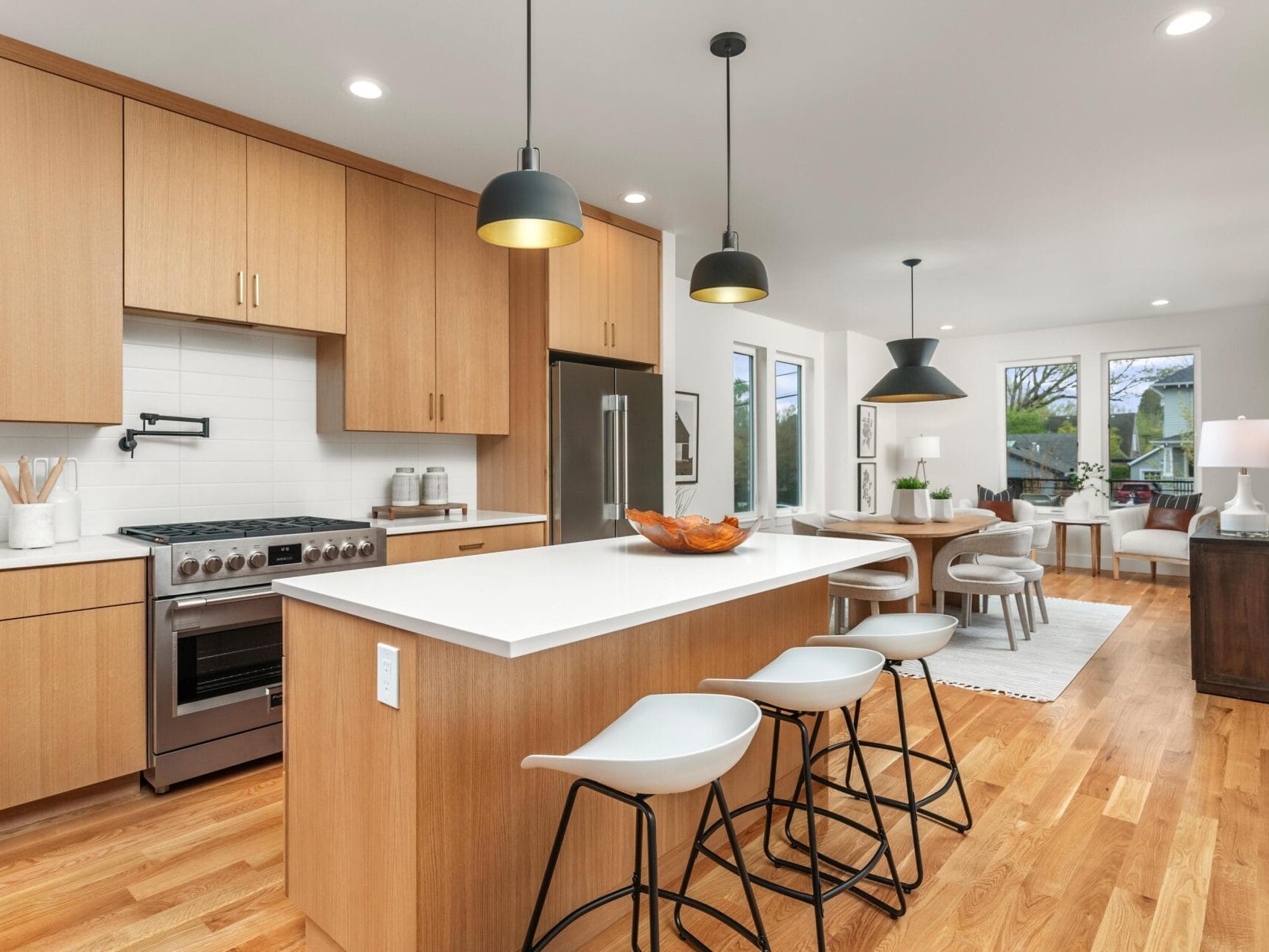 Modern kitchen and dining area with wooden cabinets, white countertops, and an island with bar stools. Pendant lights hang above the island. A dining table is set nearby, and a cozy living space is visible in the background.