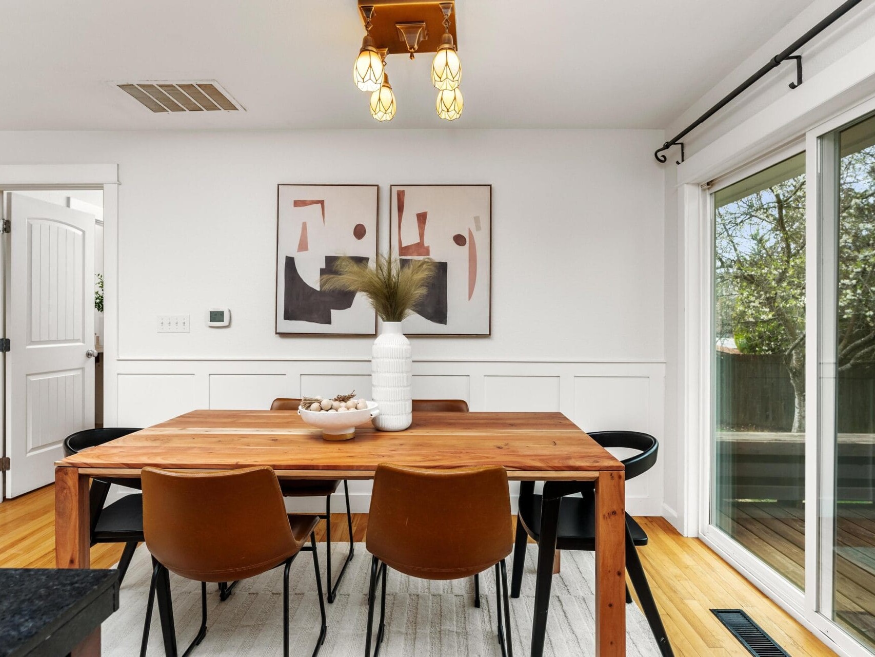 A modern dining room with a wooden table, four brown chairs, a white vase with dried grass, abstract wall art, light fixture, and large sliding glass door overlooking a deck and trees.