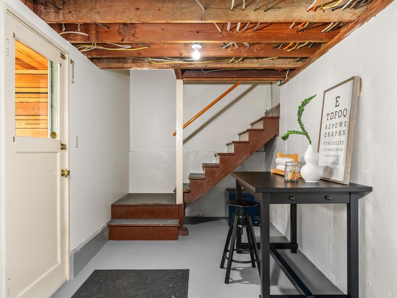 Basement with wooden stairs and a side table. Exposed beams and electrical wires on the ceiling. A chart and plant on the table, and a light above. A door and mats on the floor complete the setup.