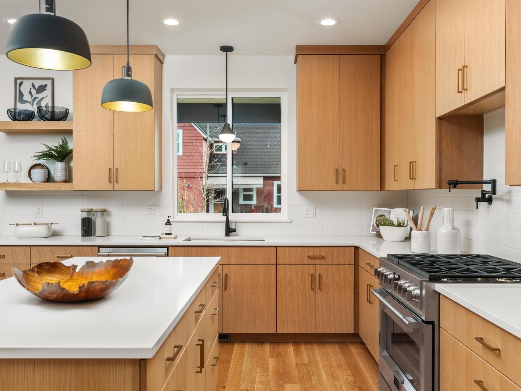 Modern kitchen with wooden cabinets, white countertops, and a central island featuring a decorative bowl. Stainless steel stove and overhead black pendant lights. A window offers a view of neighboring houses. Walls are white with minimalist decor.