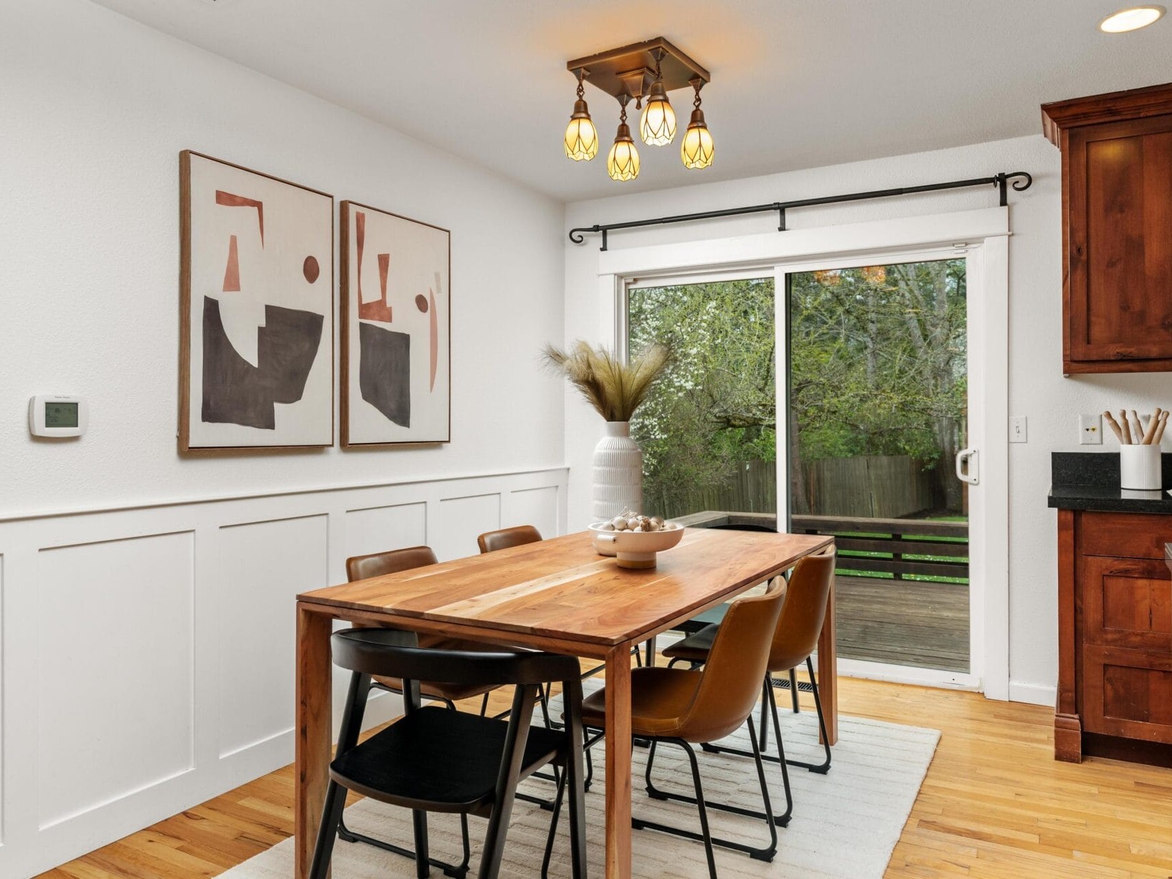 A modern dining area with a wooden table, six brown chairs, abstract art on the wall, a light fixture above, and a sliding glass door leading to a green backyard. A vase with dried plants sits on the table.