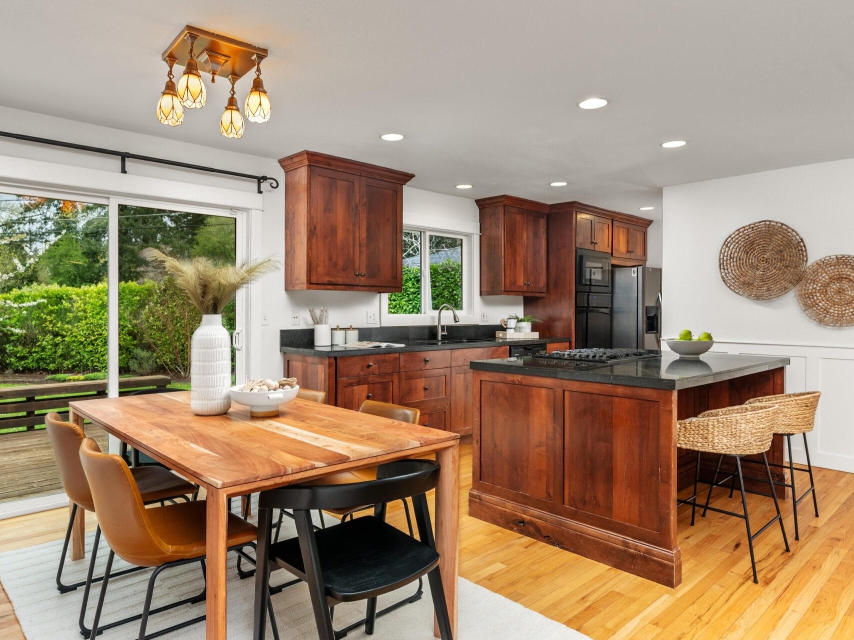 A modern kitchen and dining area with wooden cabinets, an island, black appliances, a wooden dining table with mixed chairs, large windows, and a sliding glass door overlooking a green backyard.