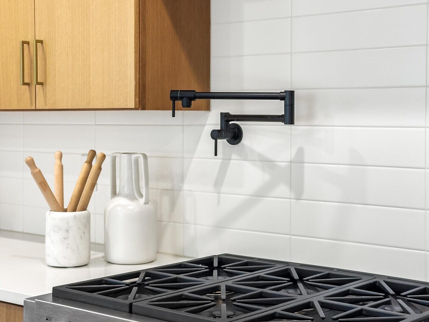 Modern kitchen with a stainless steel gas stove featuring six burners and a digital display. Theres a pot filler faucet above it, white tile backsplash, light wood cabinets, and a countertop with a white pitcher and utensil holder with rolling pins.
