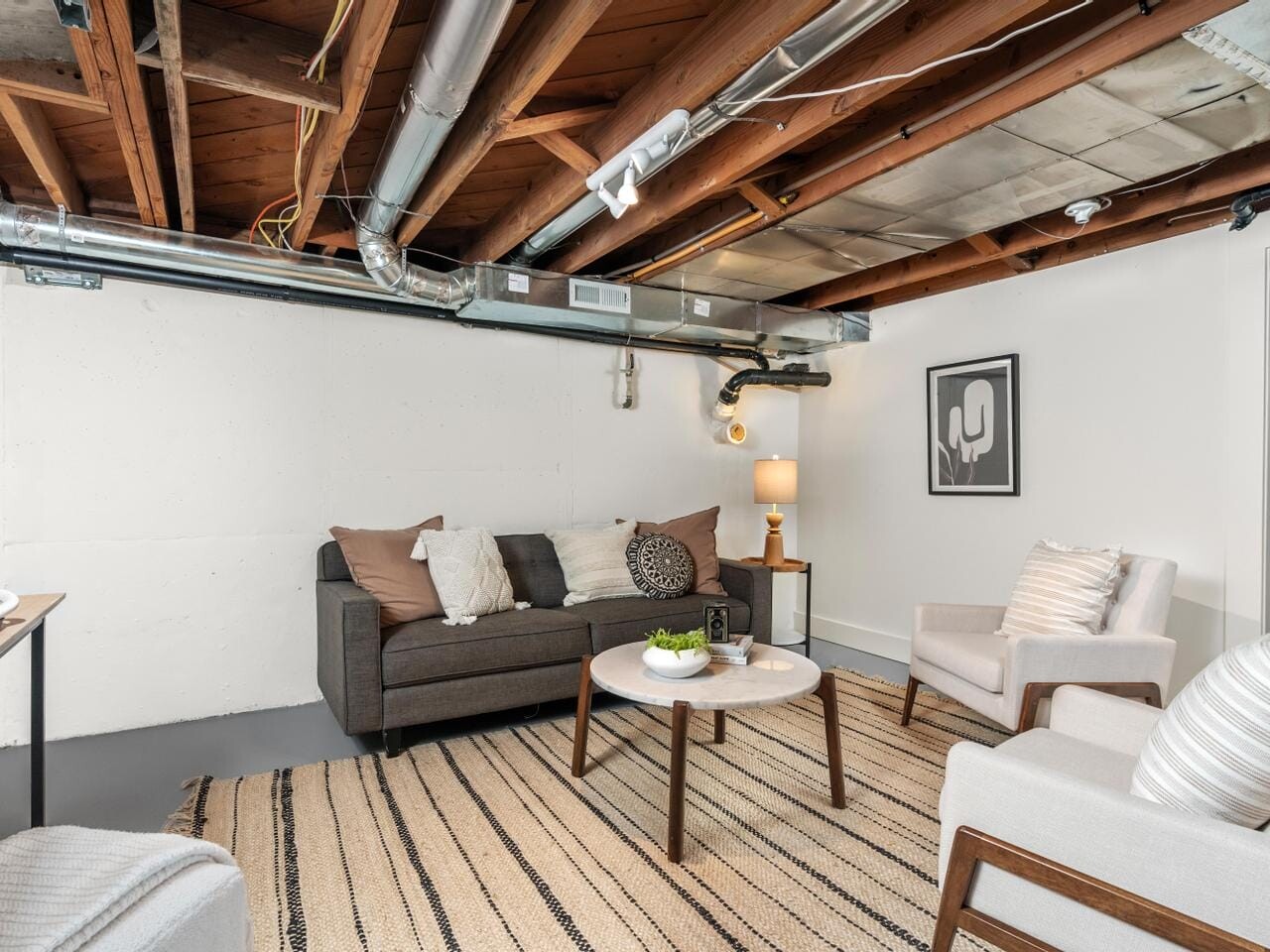 A cozy basement living area features exposed wooden beams and ductwork. It is furnished with a dark gray sofa, two light armchairs, and a round coffee table on a striped rug. A modern lamp and framed art add style to the space.
