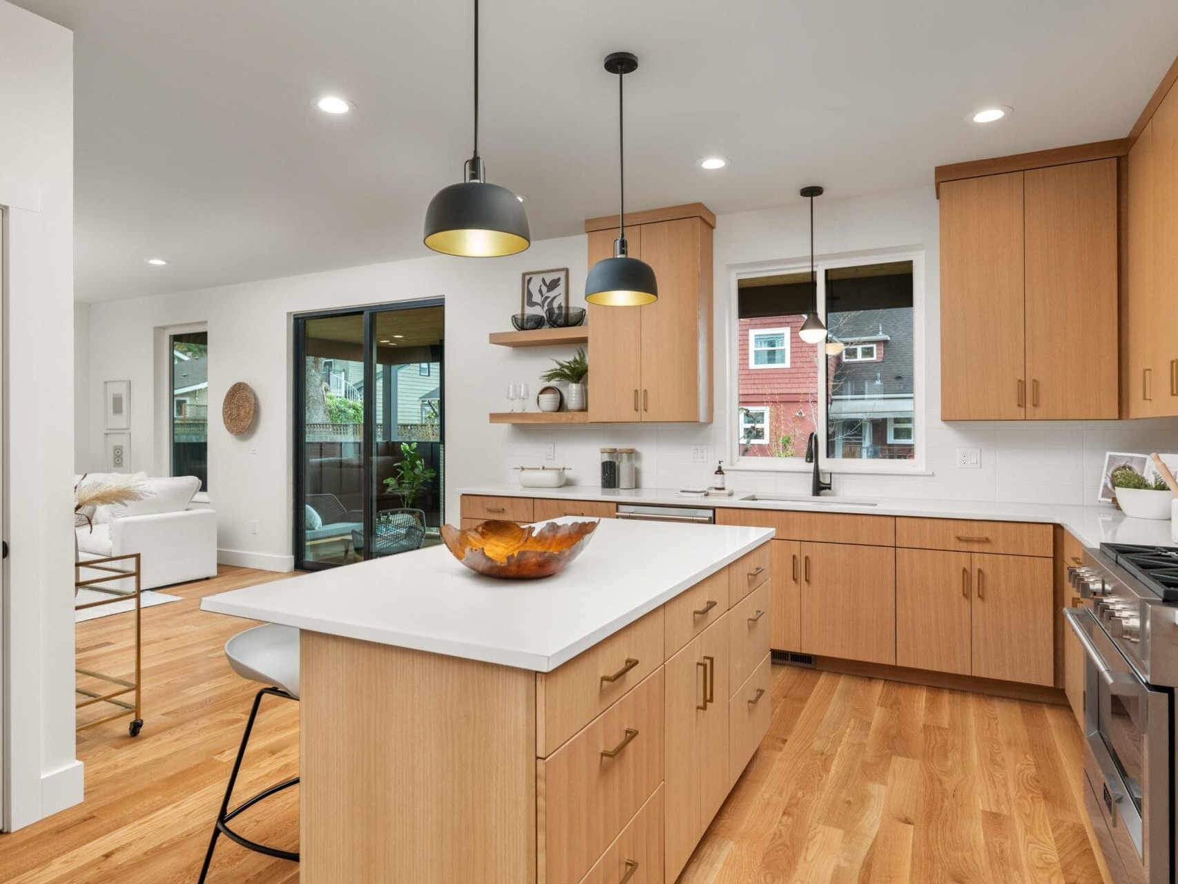 A modern kitchen with light wood cabinets and a central island. Three pendant lights hang above the island. The room features stainless steel appliances, a white backsplash, and hardwood floors. Large windows and a sliding door add natural light.