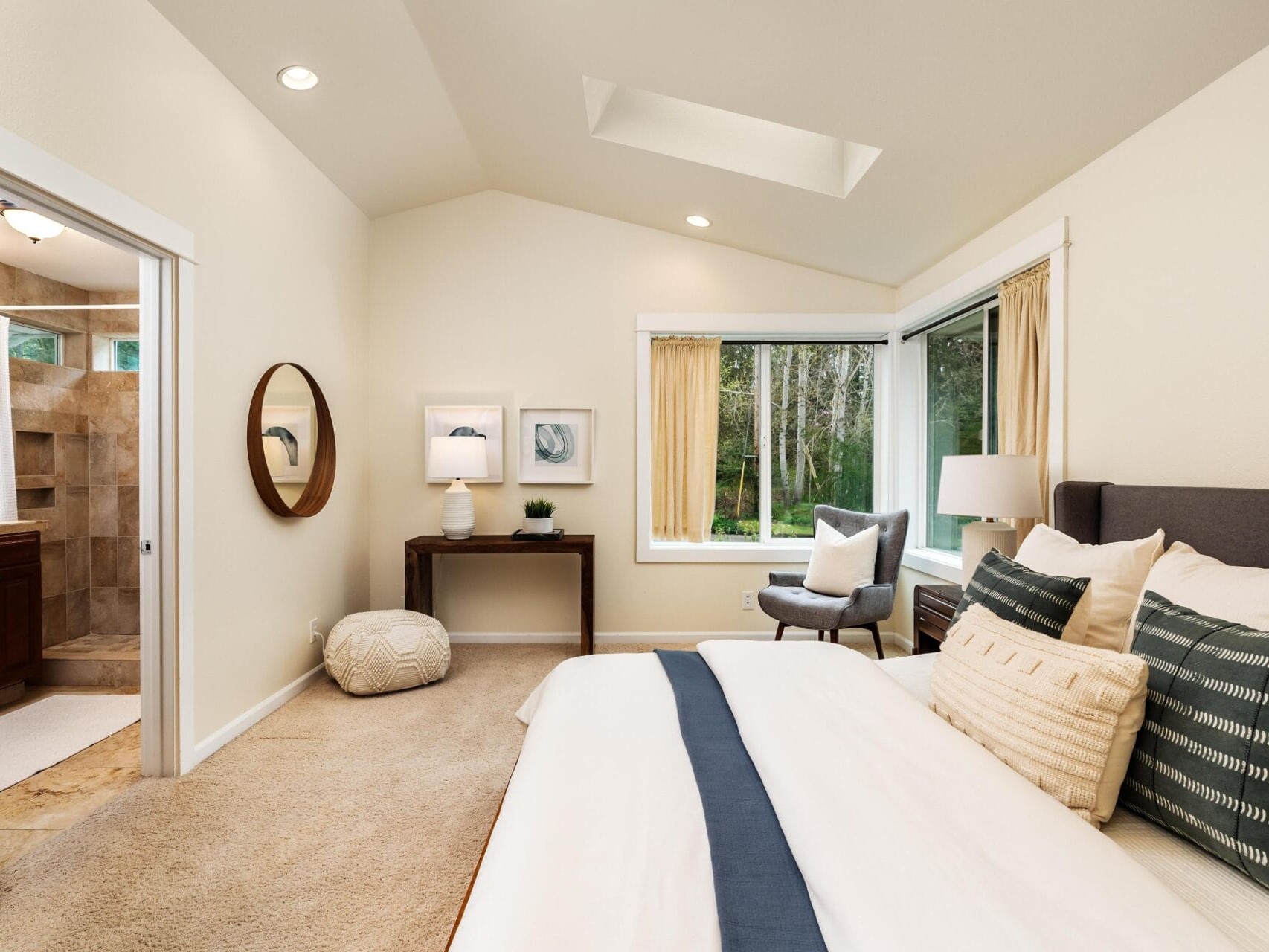 A modern, cozy bedroom with a neatly made bed, decorative pillows, a side table with lamps, a gray armchair by large corner windows, beige carpet, and an open doorway leading to a bathroom.