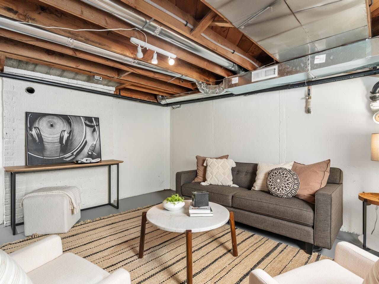Basement room with exposed wooden beams and ducts, featuring a gray sofa with mixed pillows, a striped rug, a round white coffee table, chairs, a lamp, and abstract art on a side table against a white brick wall.