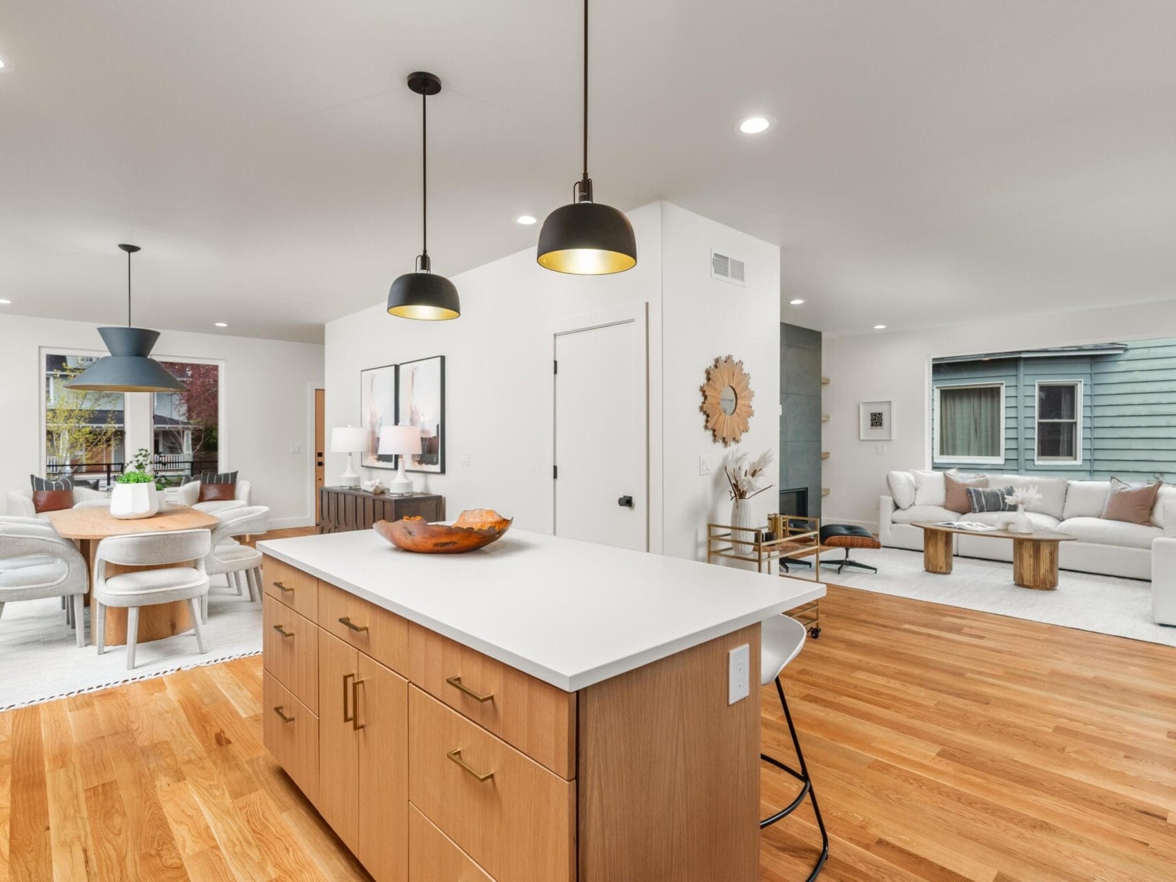 Modern open-plan home interior featuring a kitchen island with pendant lights, wooden flooring, a dining area with a round table and chairs, and a living room with a white sofa and armchairs. Light streams in through large windows.