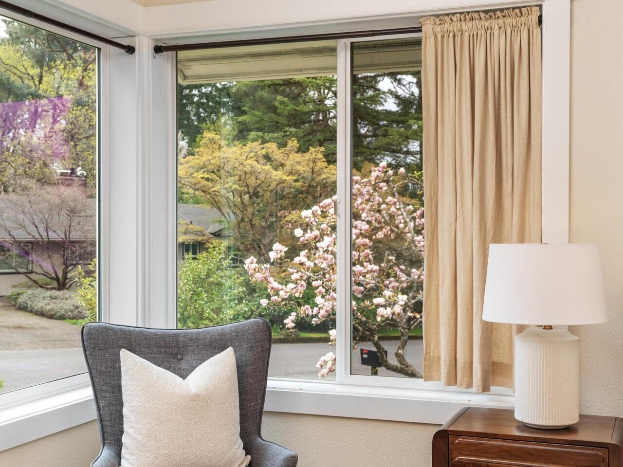A cozy corner with a gray armchair and white pillow sits by large windows overlooking blooming trees. A wooden side table with a lamp and basket is beside the chair on a beige carpet.