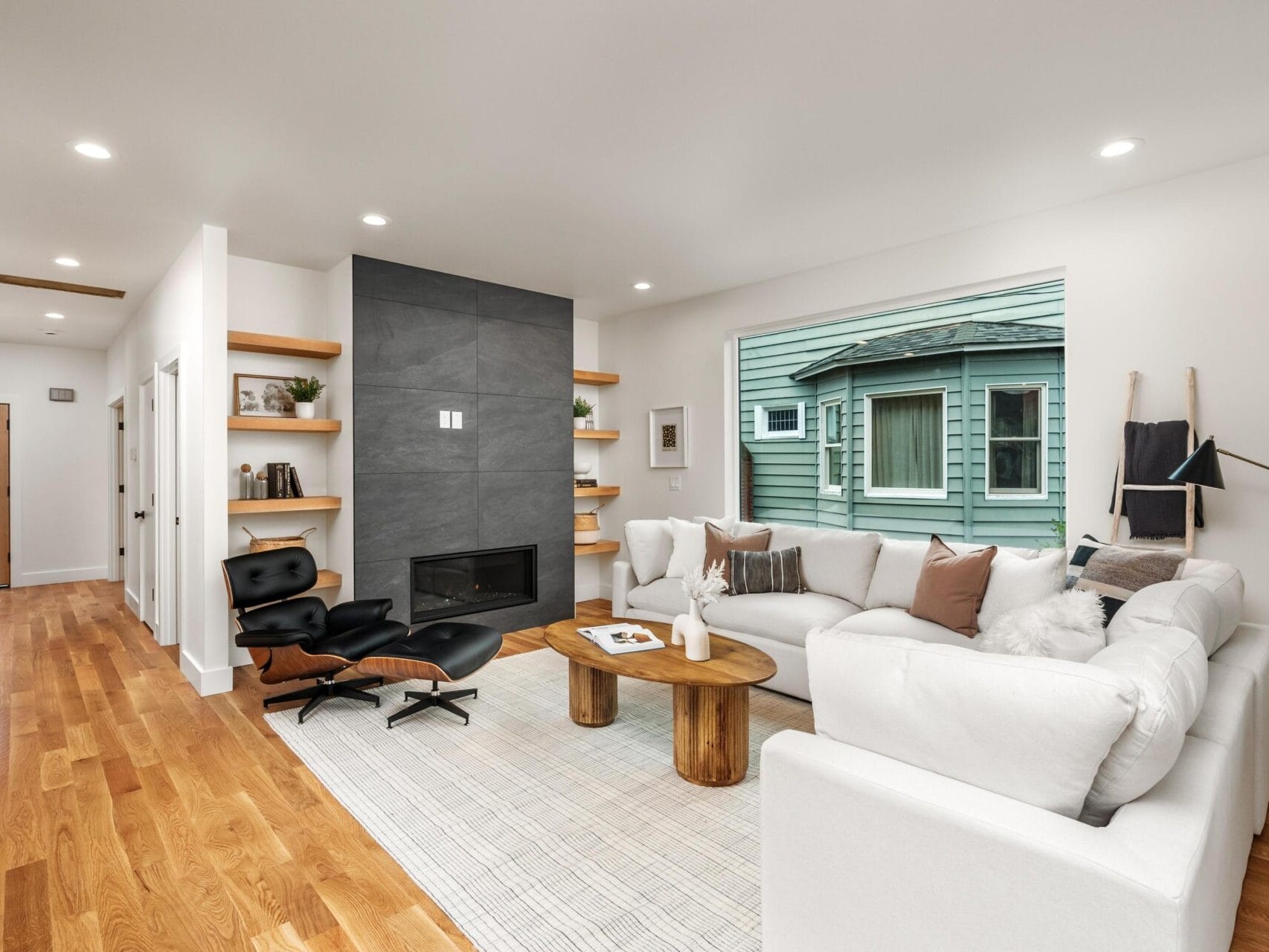 Modern living room with a large window, white walls, and wood flooring. Features a white sectional sofa, black lounge chair, minimalist coffee table, built-in shelves, and a dark accent wall with a fireplace. Outside view of a neighboring house.