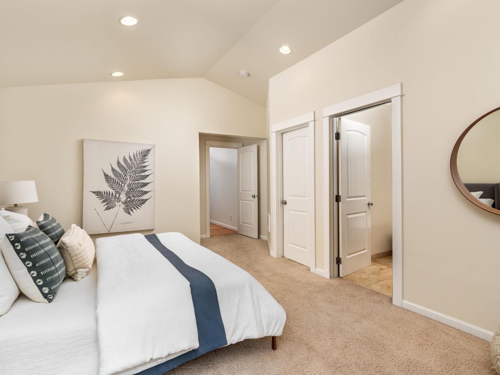 A cozy bedroom with a neatly made bed featuring white linens and blue accents, a large fern artwork on the wall, two white doors, a round mirror, and soft beige carpeting under warm recessed lighting.