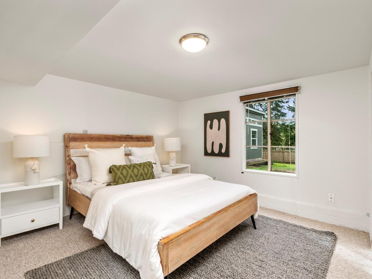 A cozy bedroom features a wooden bed with white bedding and green accent pillow, flanked by two white nightstands with lamps. A modern art piece hangs above. A window offers a view of a grassy yard and trees. The room is carpeted in gray.