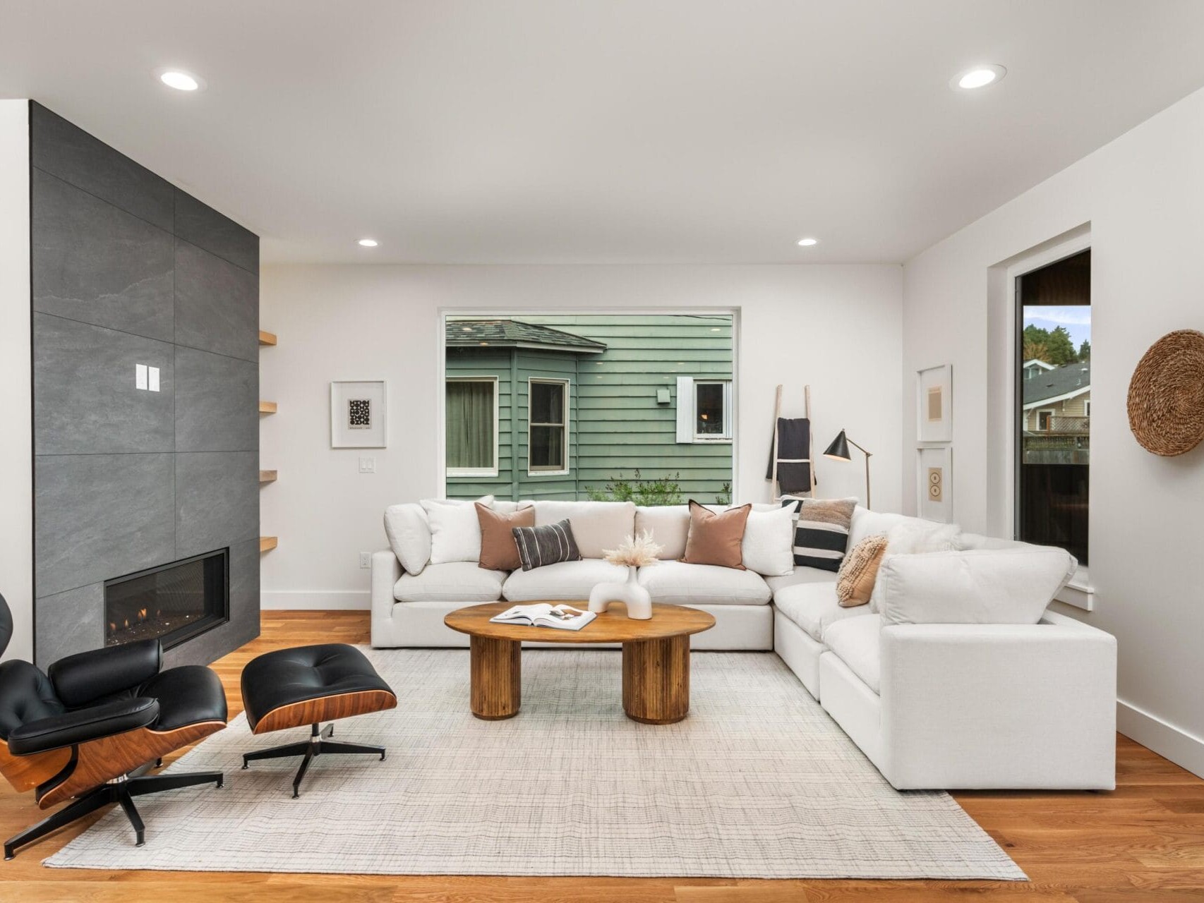 Modern living room with a large white sectional sofa, wooden coffee table, and leather lounge chair. Minimalist decor includes a wall-mounted fireplace, floating shelves, and a large window. Neutral tones and wooden flooring add warmth to the space.