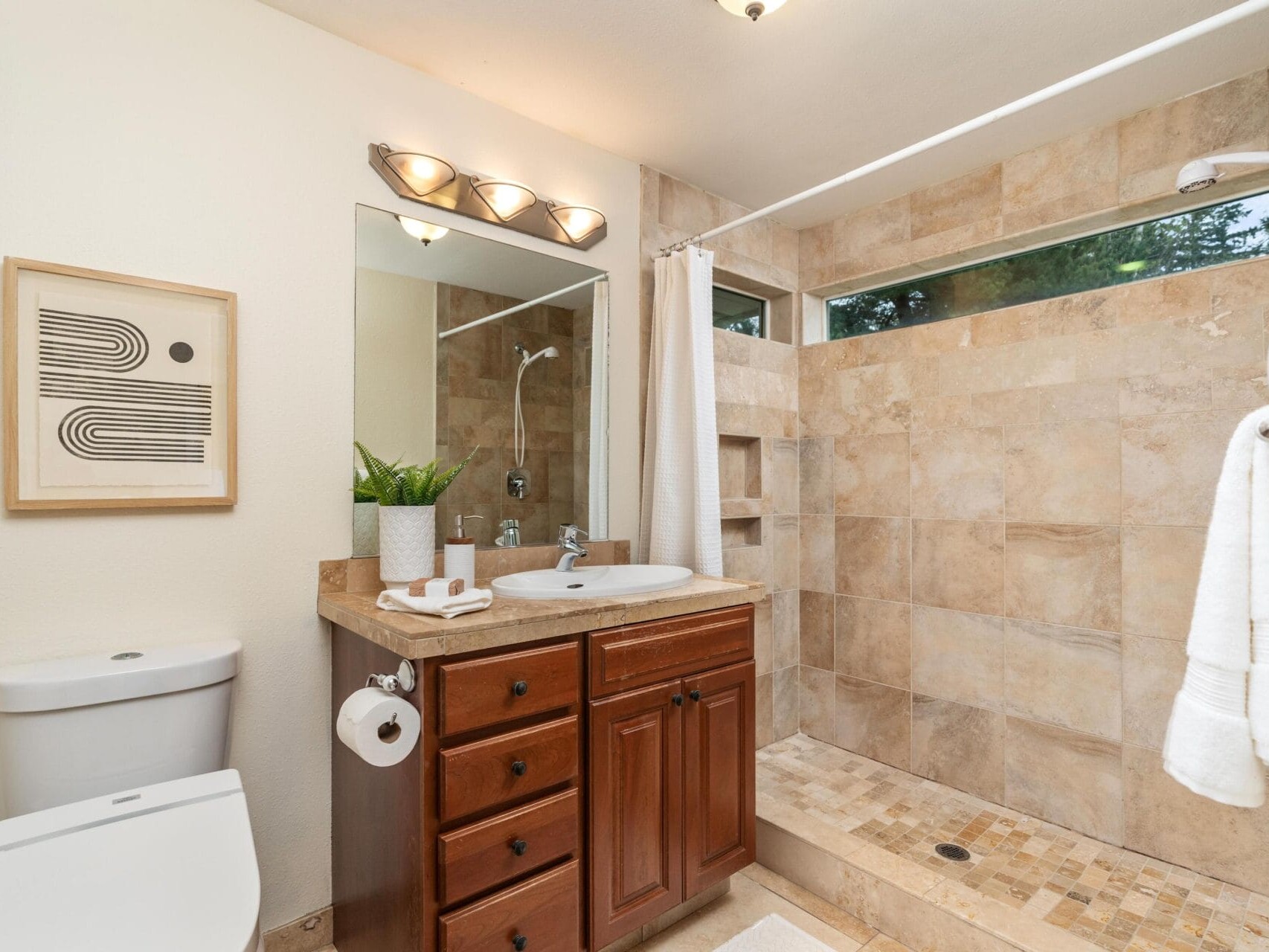 A modern bathroom with a large walk-in tiled shower, a wooden vanity with a sink, a toilet, a potted plant, a wall mirror, towel racks with white towels, and abstract wall art.