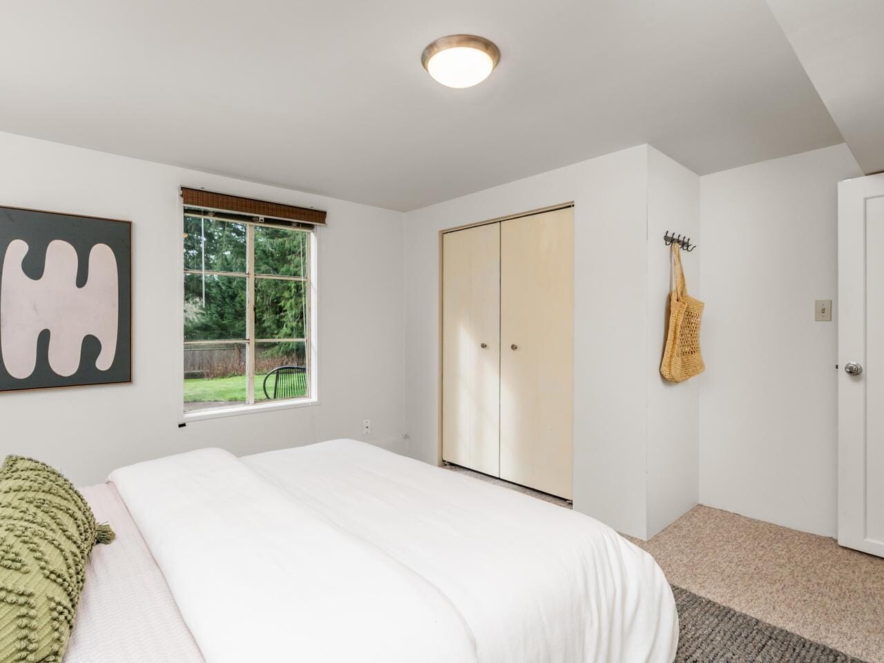 A minimalist bedroom with a large bed featuring green and white pillows, soft bedding, and a bedside table. The walls are white, with a modern abstract artwork. There is a window, a closet with wooden doors, and a hanging jute tote bag.