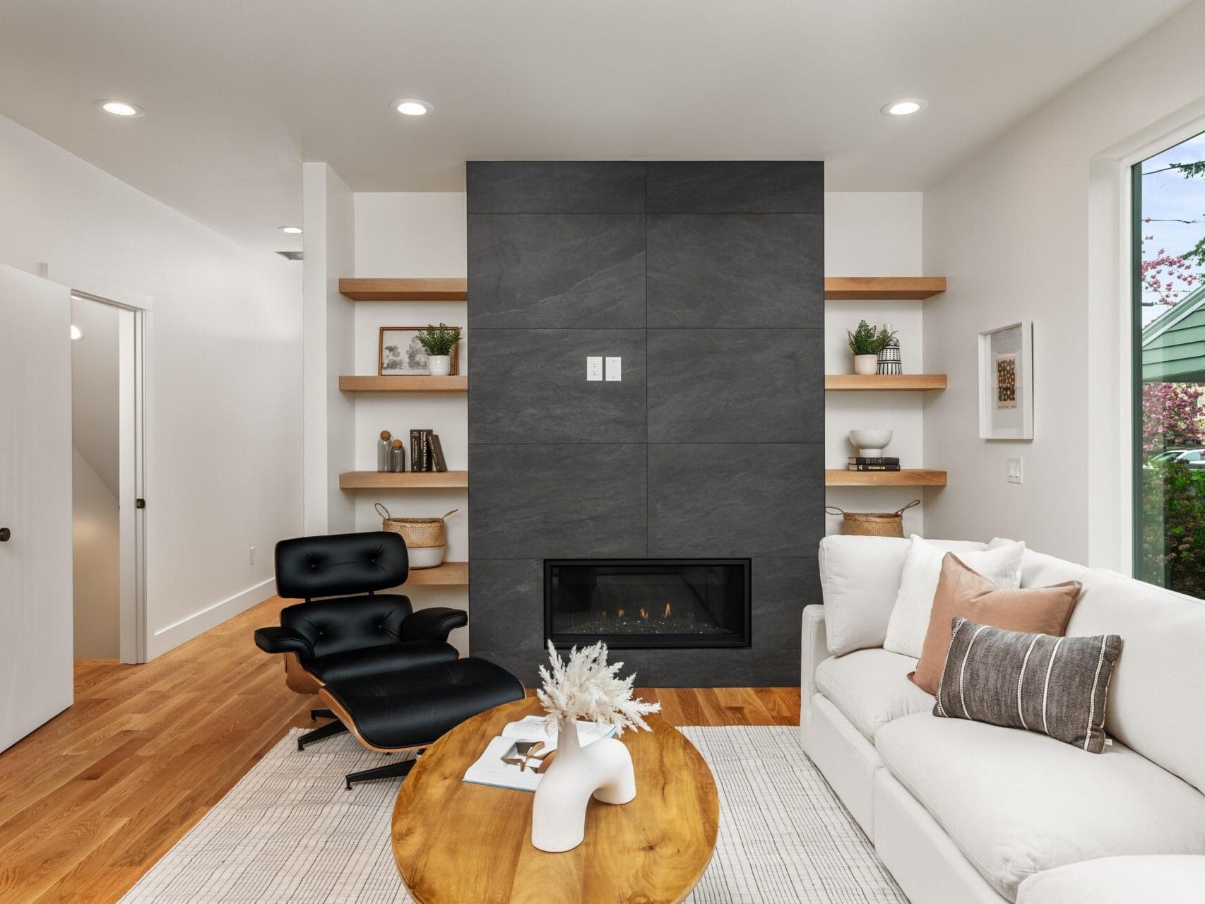 Modern living room with a gray accent wall featuring a fireplace, flanked by wooden shelves with decor. A white sofa and a black lounge chair with an ottoman face a wooden coffee table. Large window shows greenery outside.