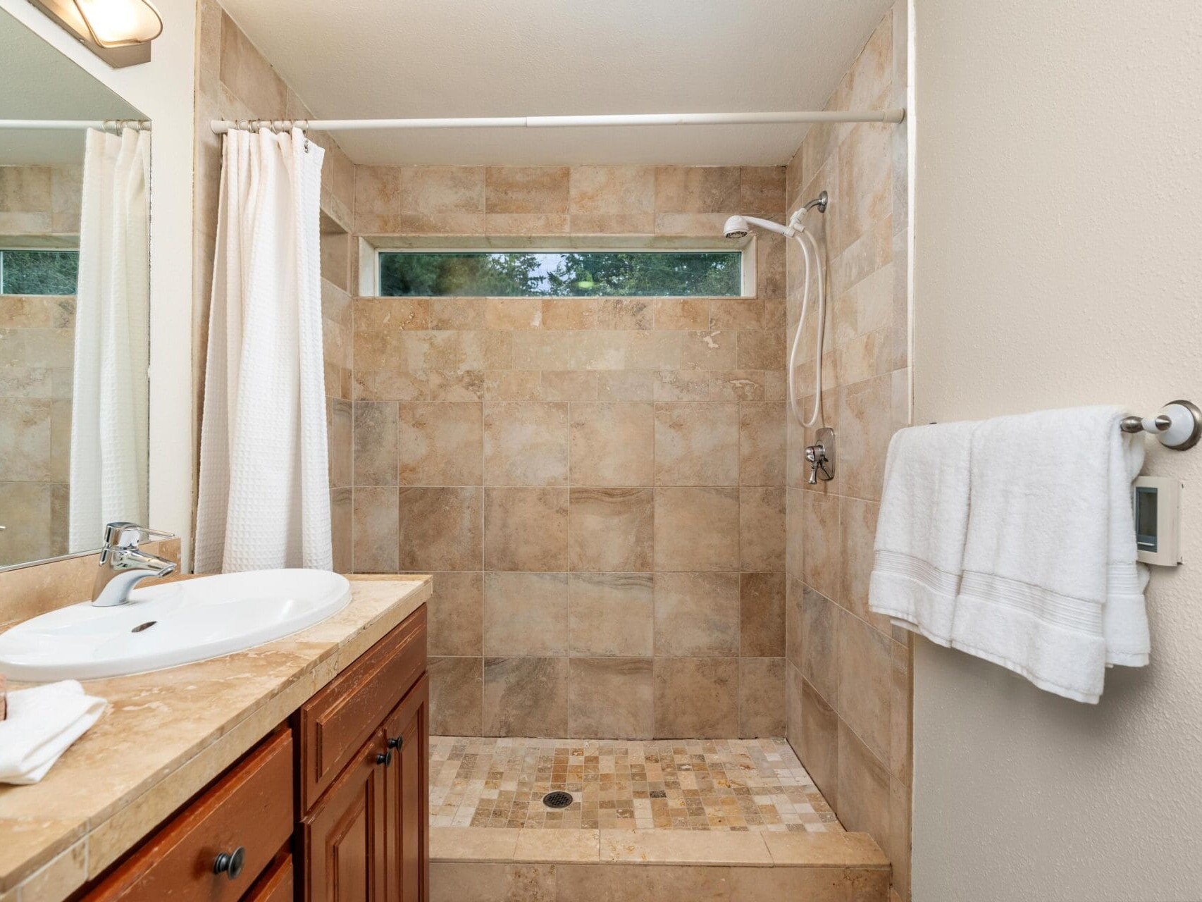 A bathroom with a tiled walk-in shower, a white curtain, a sink with a wooden vanity, a mirror, and a towel hanging on a rack on the wall. There is a horizontal window in the shower area.